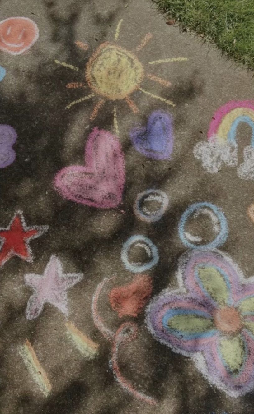Colorful chalk drawings on a concrete sidewalk including a smiling sun, hearts, stars, rainbows, butterflies, and circles.