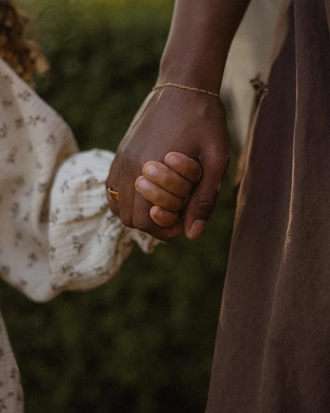 A close-up of two people holding hands, the person on the right is wearing a brown sleeve and a gold bracelet. The person on the left is wearing a white outfit with a floral pattern.