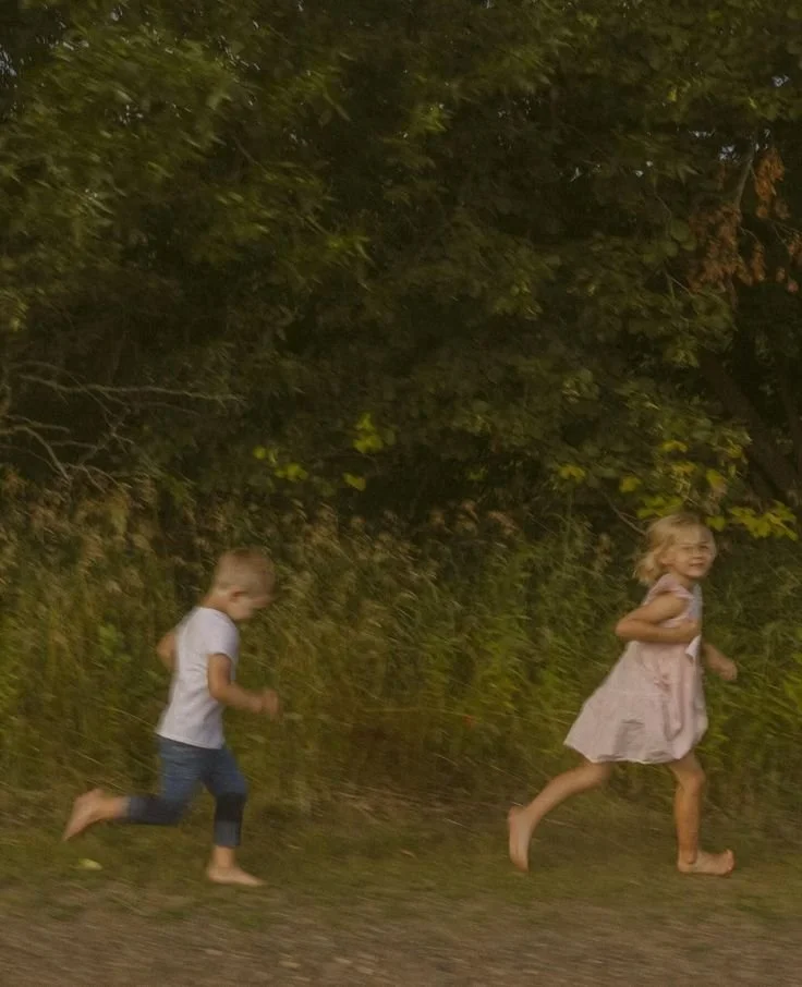 Two young children, a girl in a light pink dress and a boy in a white t-shirt and jeans, running outdoors on a dirt path with trees and bushes in the background.