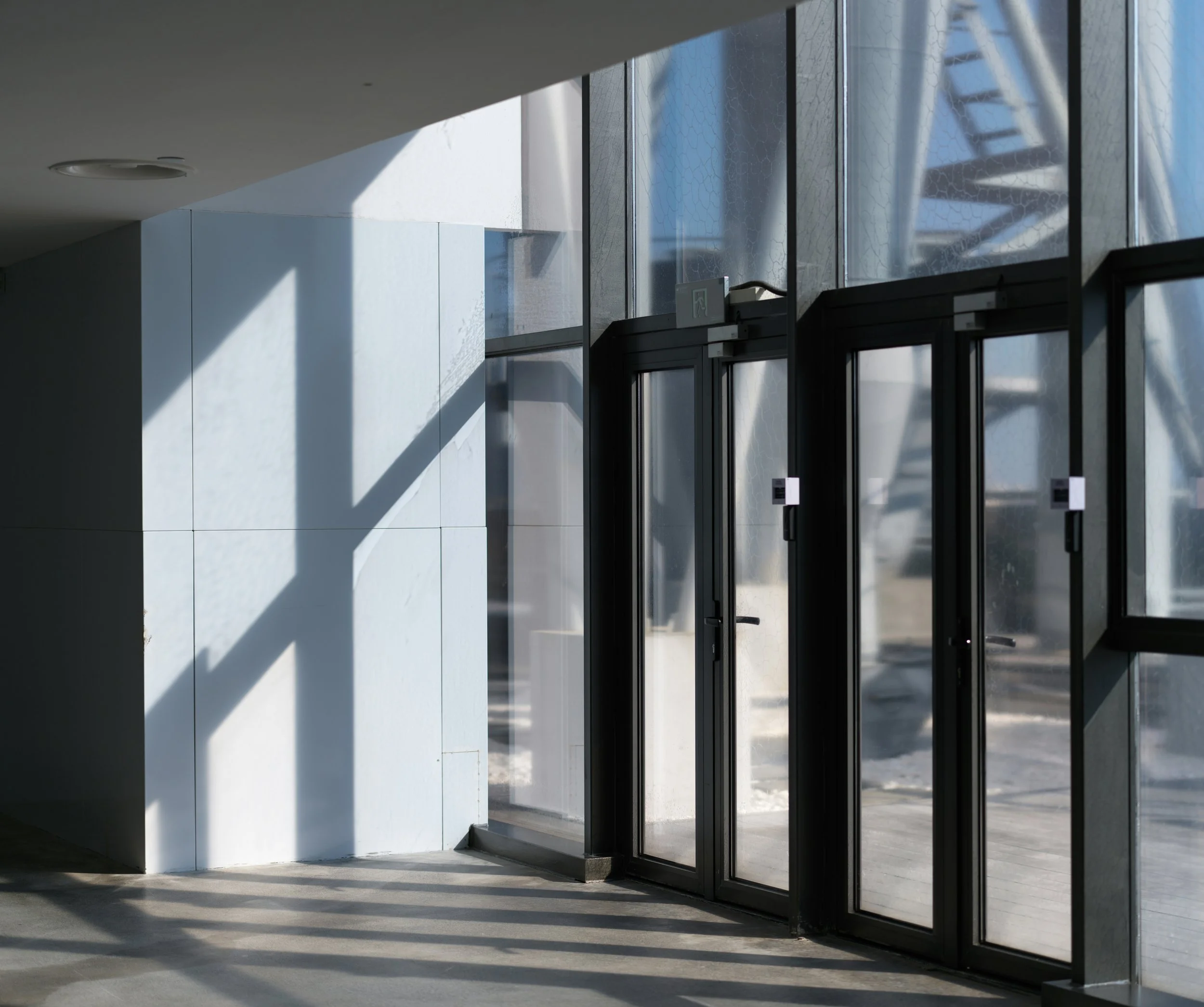 Modern building entrance with glass doors and white exterior wall, shadow cast on the ground from the sunlight.