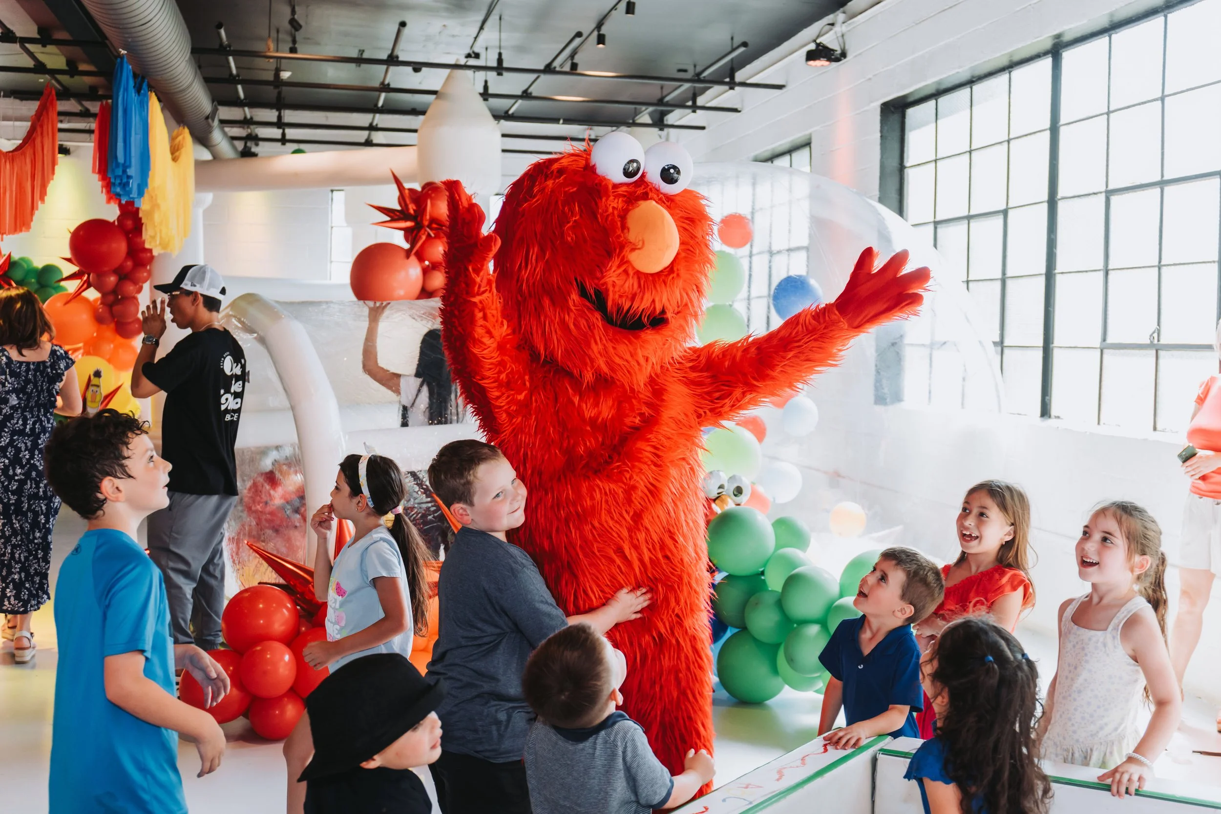 Children gathered around a person in a large, furry, red Elmo costume at a colorful party or event with balloons and decorations.