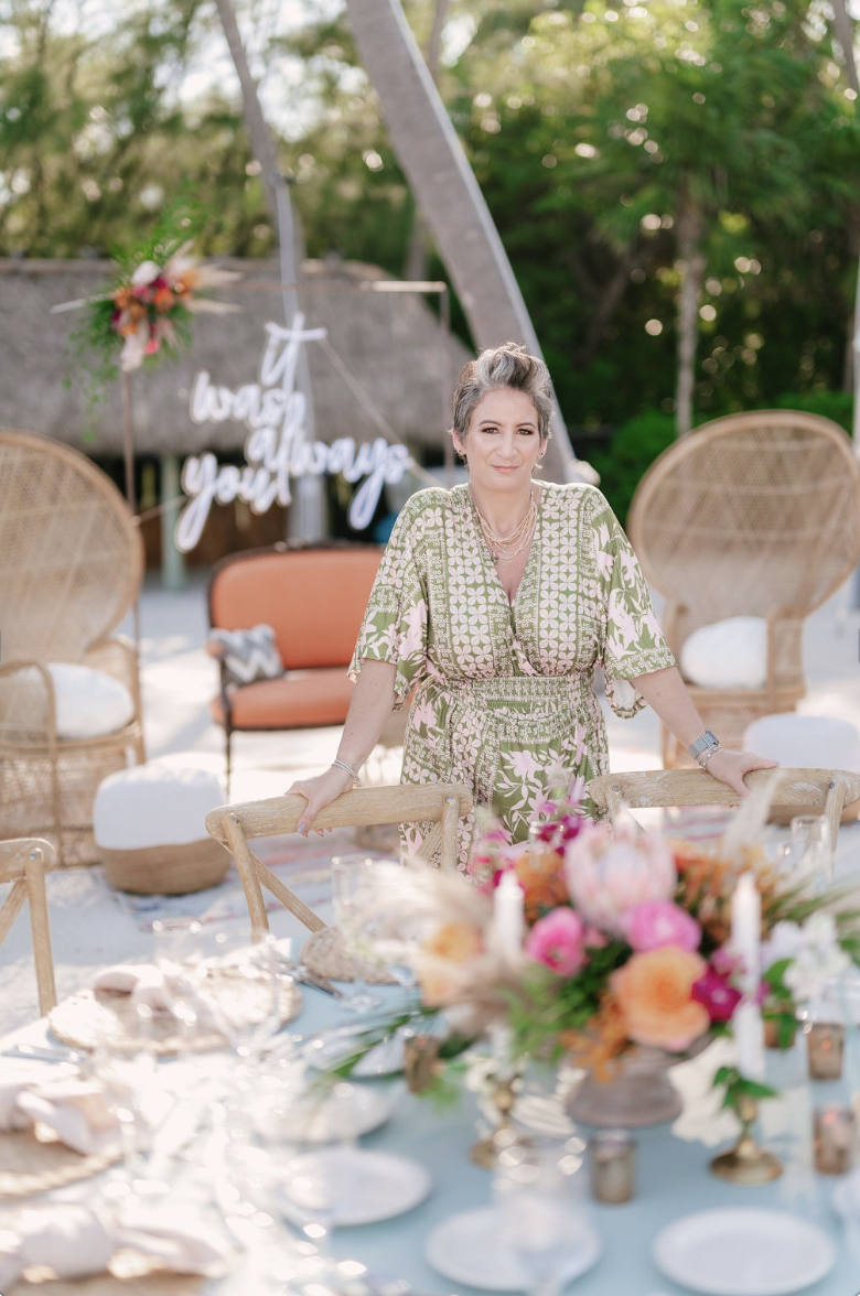 A woman with short gray hair standing behind a dining table at an outdoor event, with a backdrop decorated with a floral arrangement and a sign that says "Love You" in cursive. The setting includes wicker chairs, a floral centerpiece, and trees in the background.