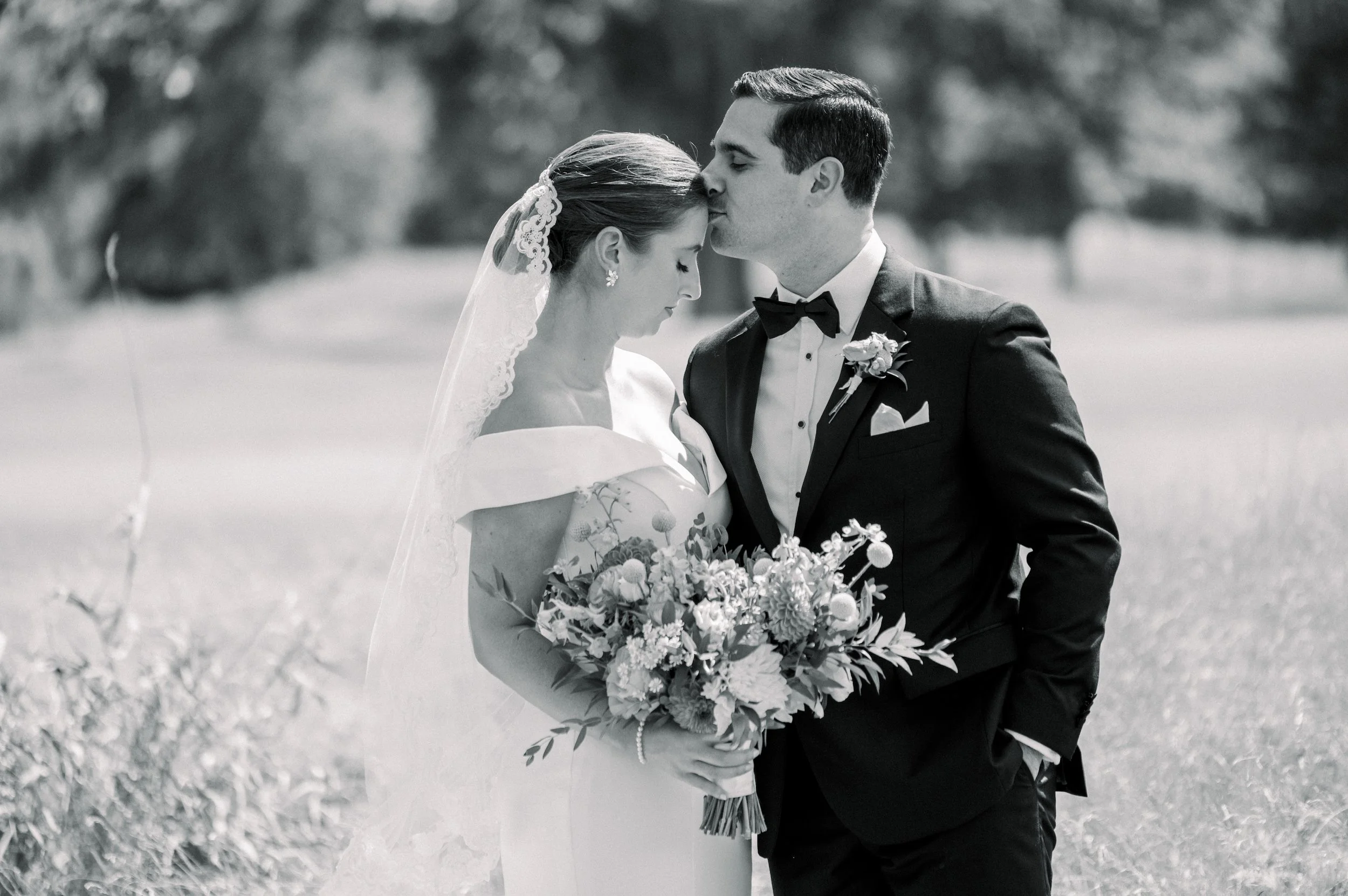A black and white photo of a bride and groom embracing outdoors, with the groom kissing the bride's forehead. The bride is holding a large bouquet of flowers and is wearing a veil and wedding dress. The groom is dressed in a tuxedo with a bow tie and