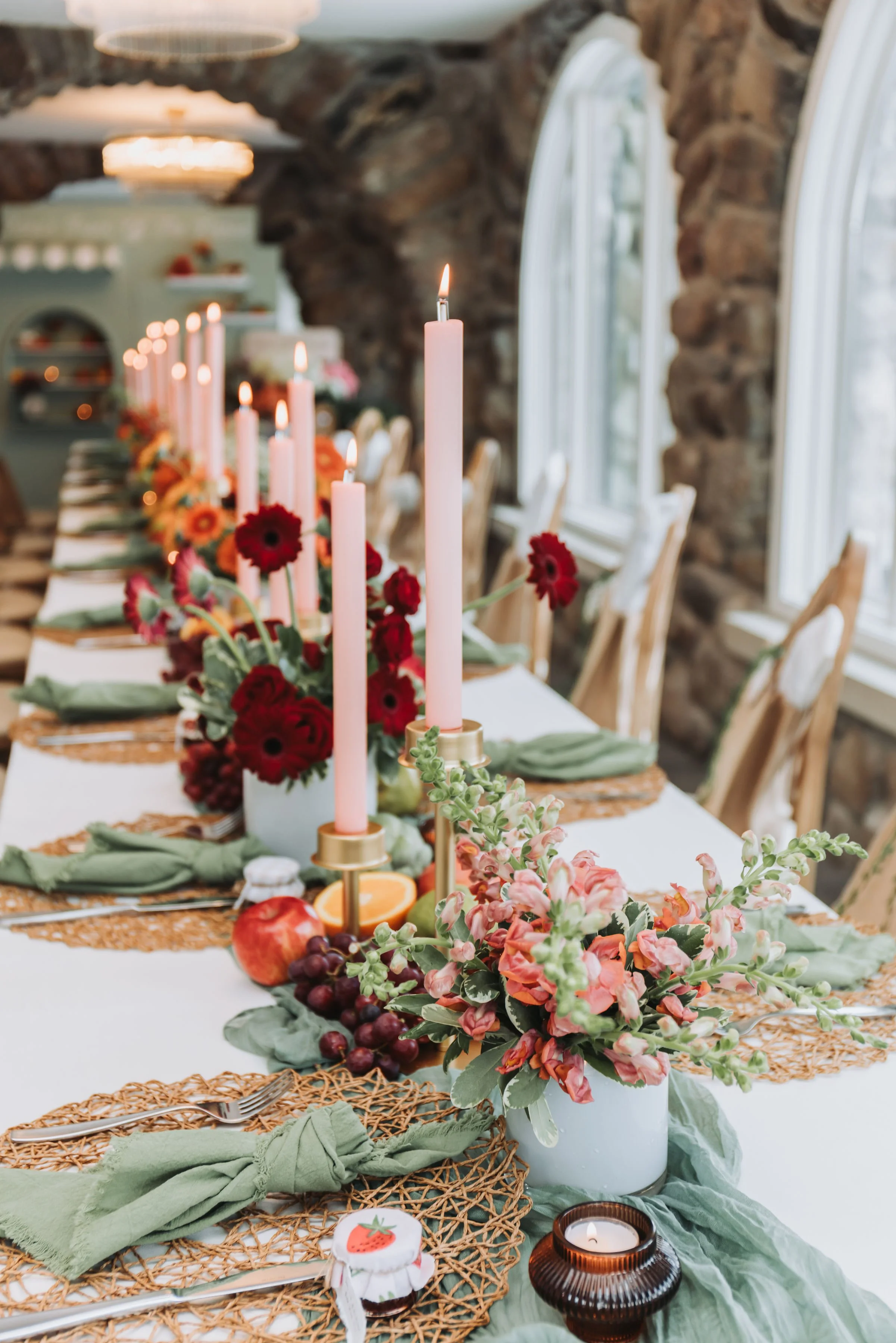 Elegant dining table set for a celebration with pink candles, floral arrangements, and green napkins, in a rustic room with stone walls and arched windows.