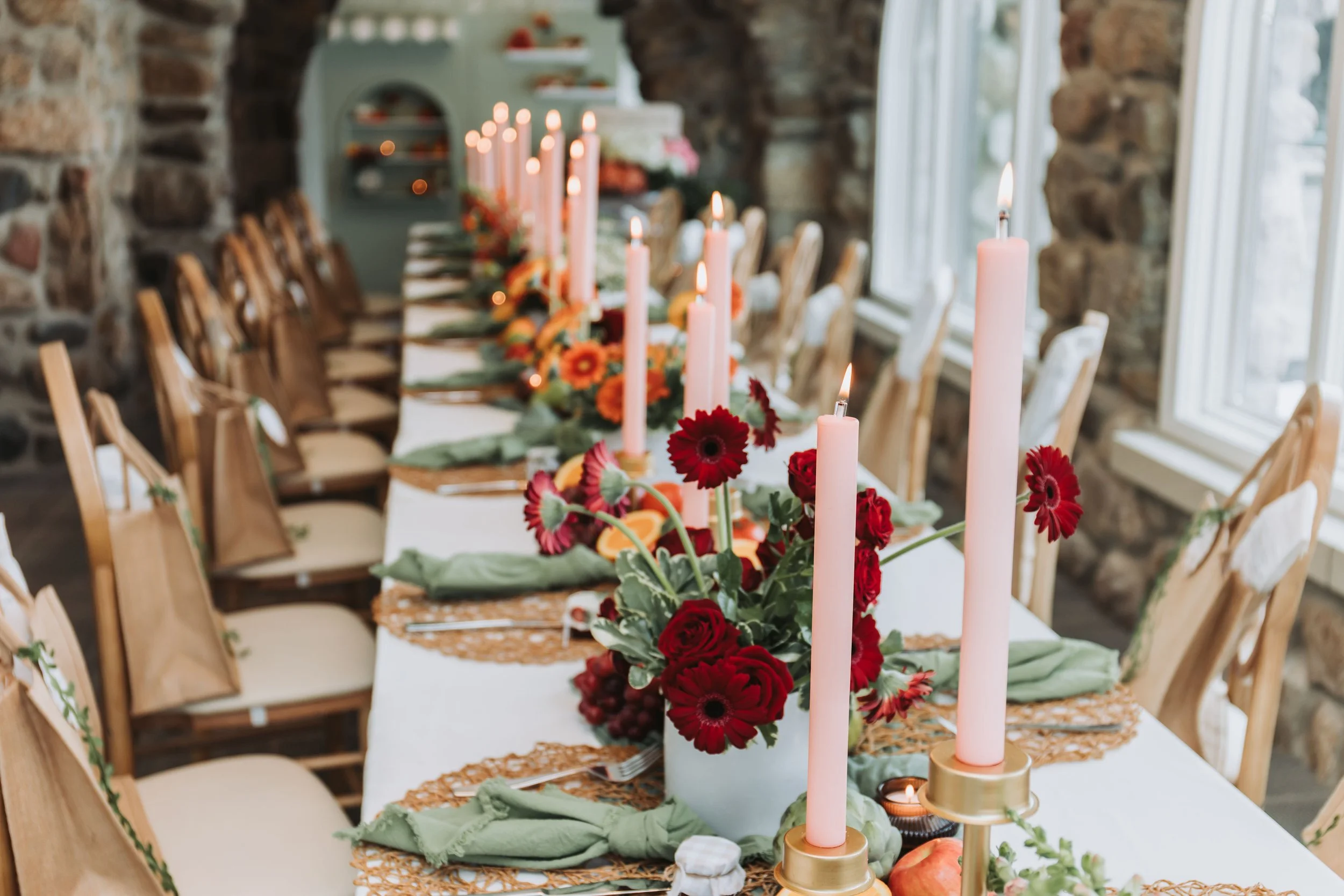 Elegant dining table decorated with pink candles, red flowers, and fall-themed accents in a rustic stone-walled room with large windows.