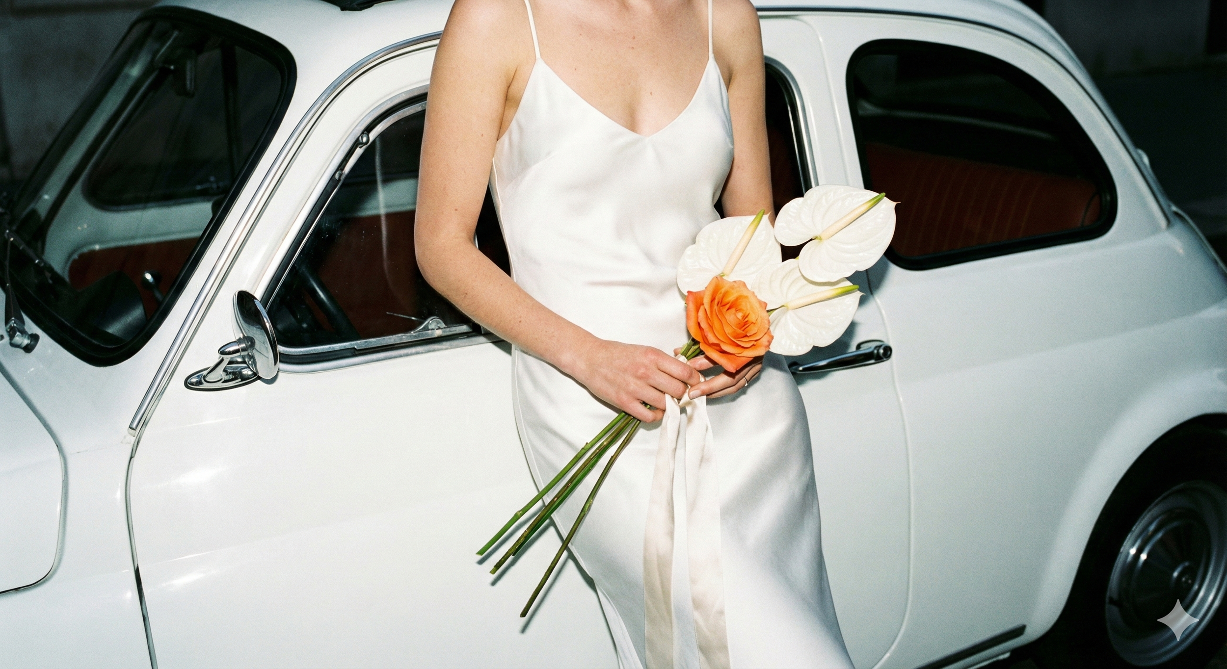 A person in a white dress holding a bouquet of orange and white flowers standing next to a white vintage car.