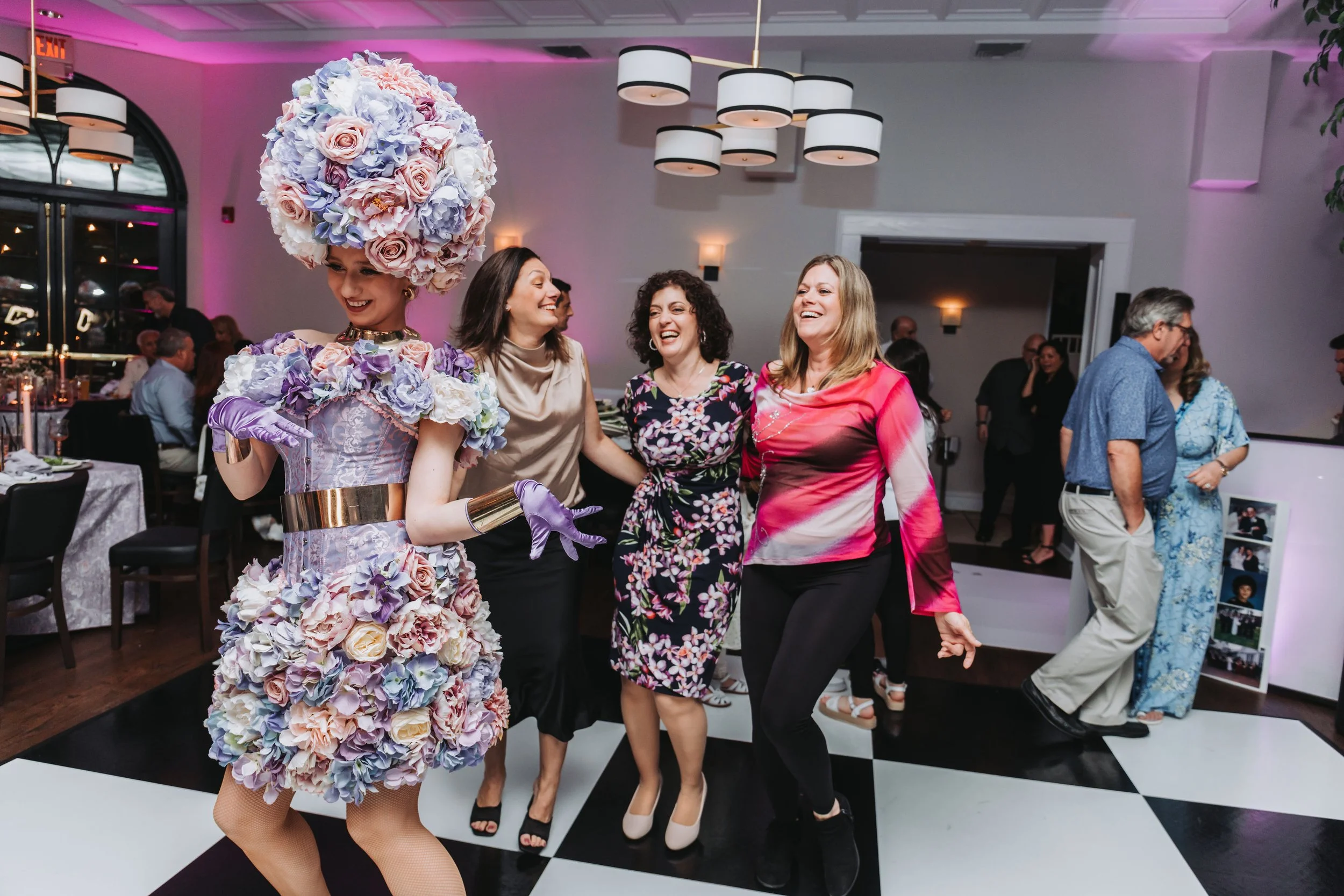 Woman dressed as a floral dress and headpiece, smiling and dancing with a group of women at an indoor celebration or party.
