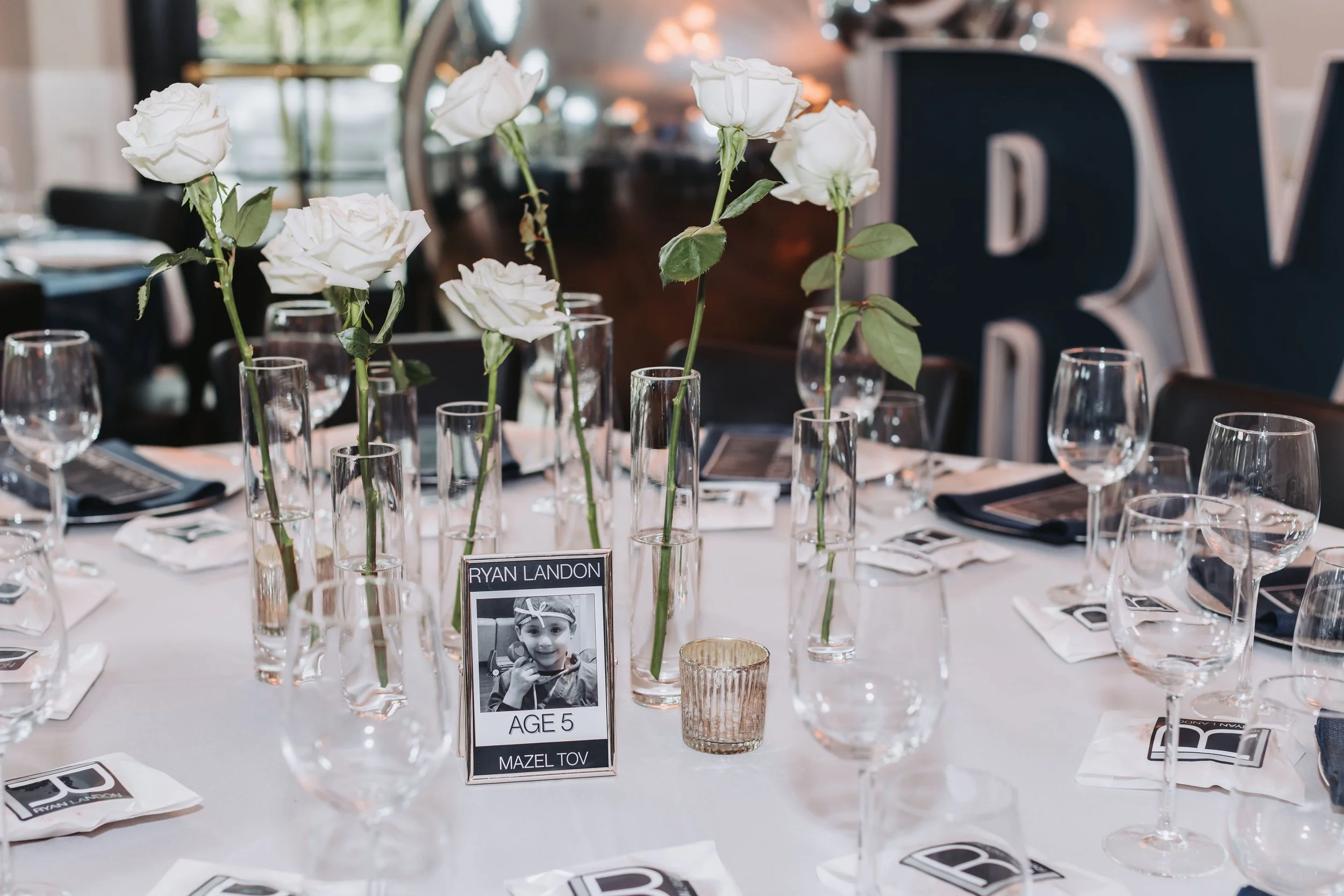 A banquet table decorated with white roses in tall vases, glasses, and place settings. A small black and white photo of a child with the name tag "Ryan Landon" and age 5 is visible on the table.