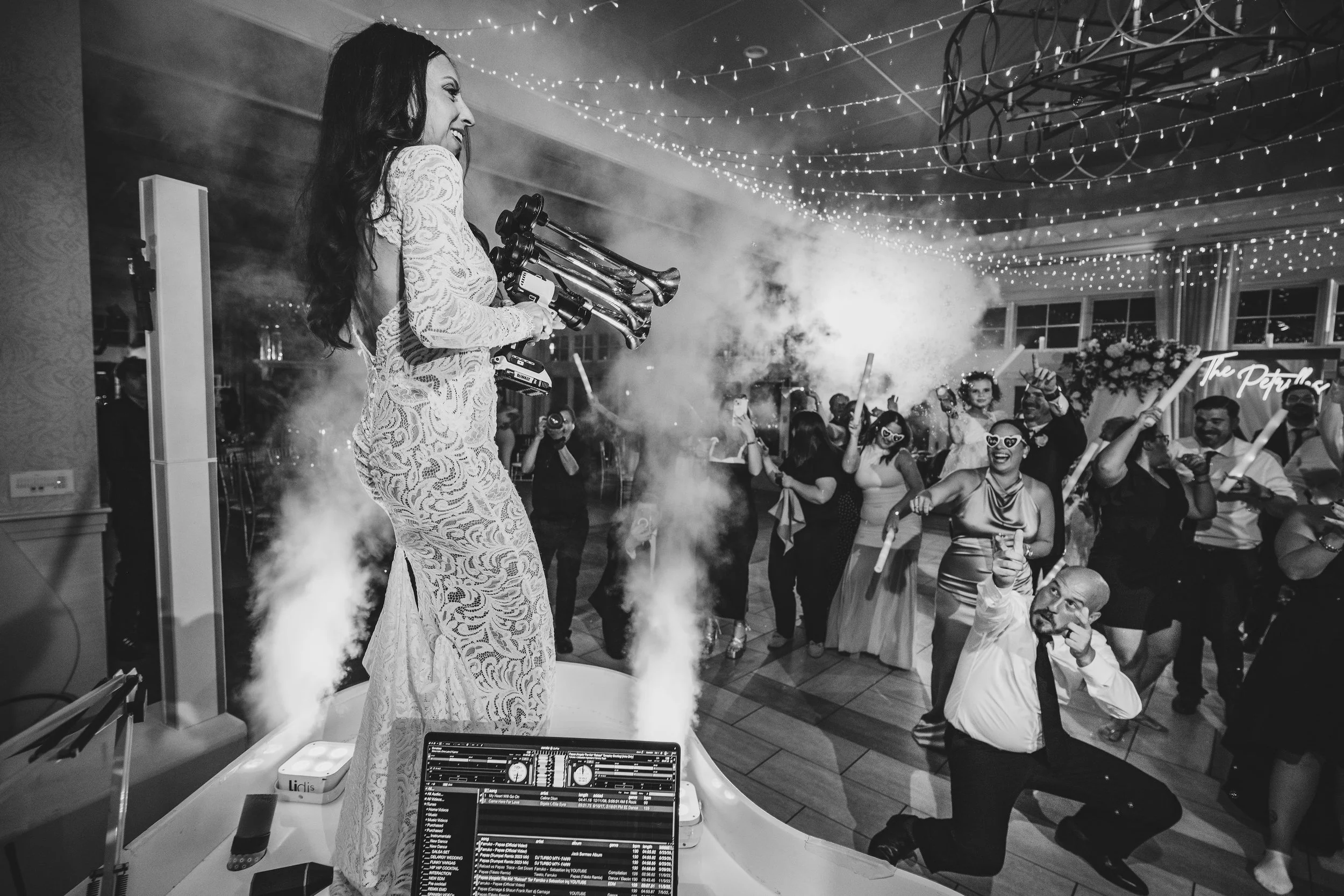 A woman in a lace dress DJing at a party with people dancing and celebrating indoors decorated with string lights.