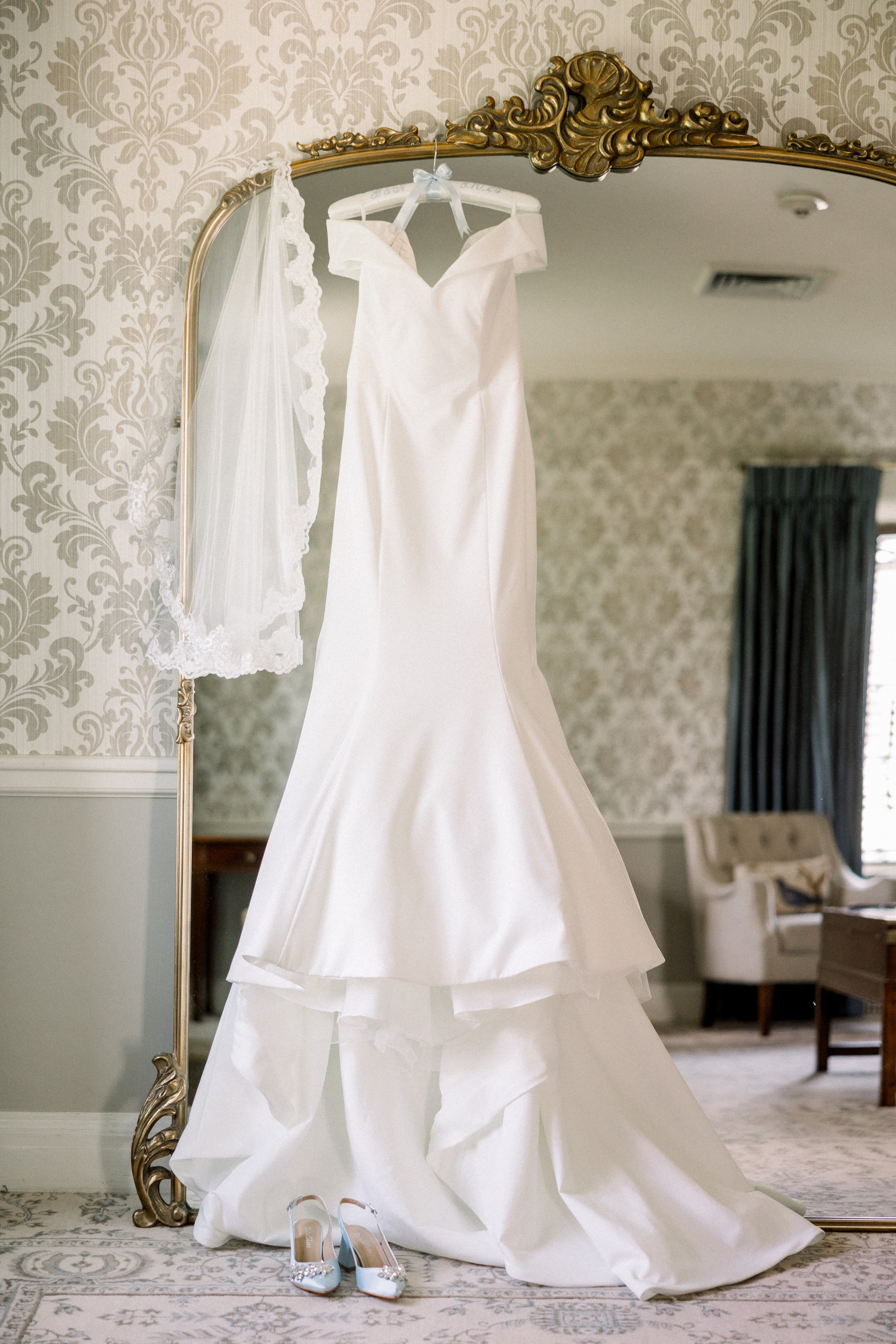 A white wedding dress is hanging on a decorative mirror with gold accents in a well-lit room. A white veil with lace trim and a pair of embellished white high-heeled shoes are on the floor in front of the mirror.