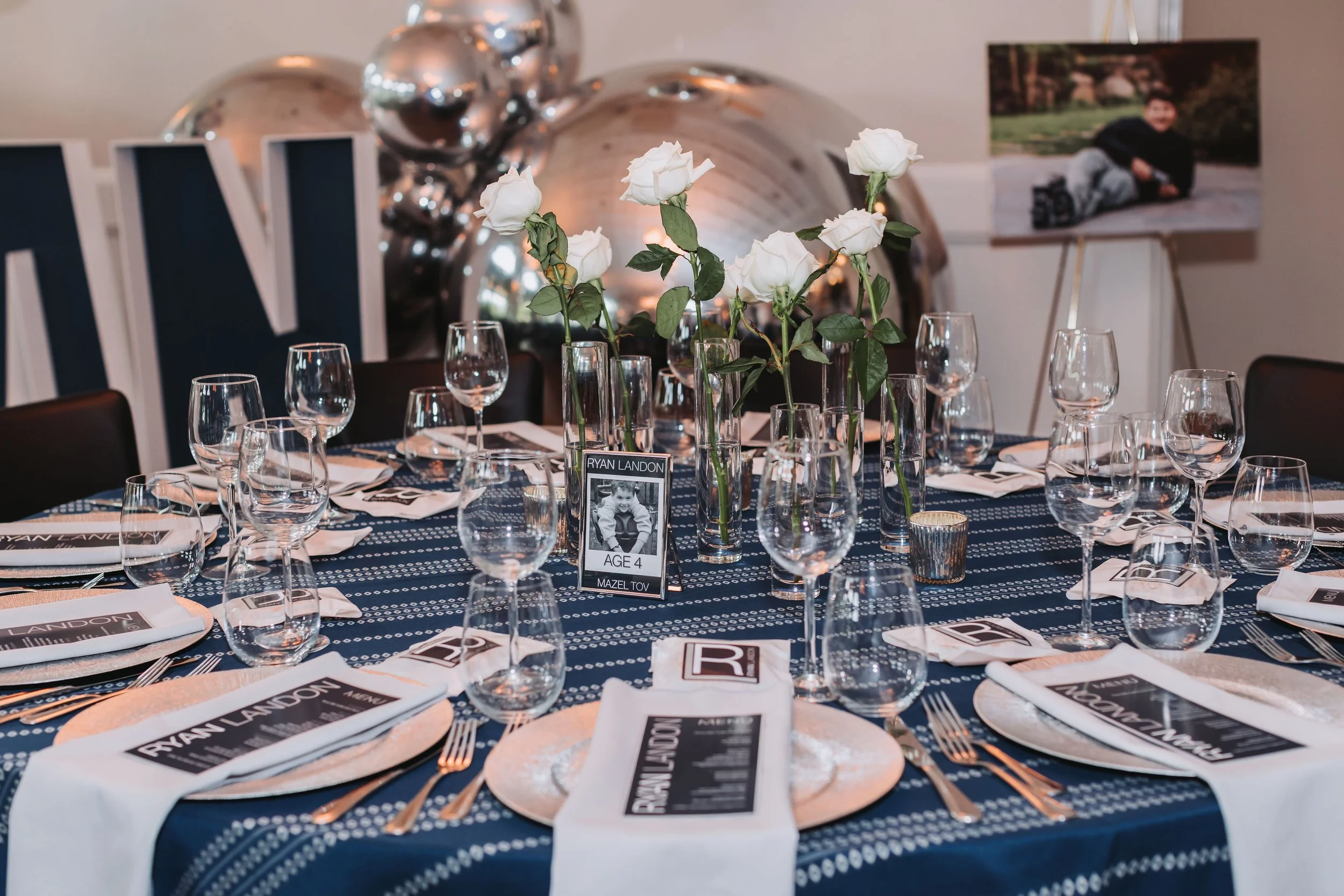 Decorated dining table with white roses in glass vases, multiple wine glasses, plates with napkins, utensils, and printed menus or invitations, set for a formal event or celebration.