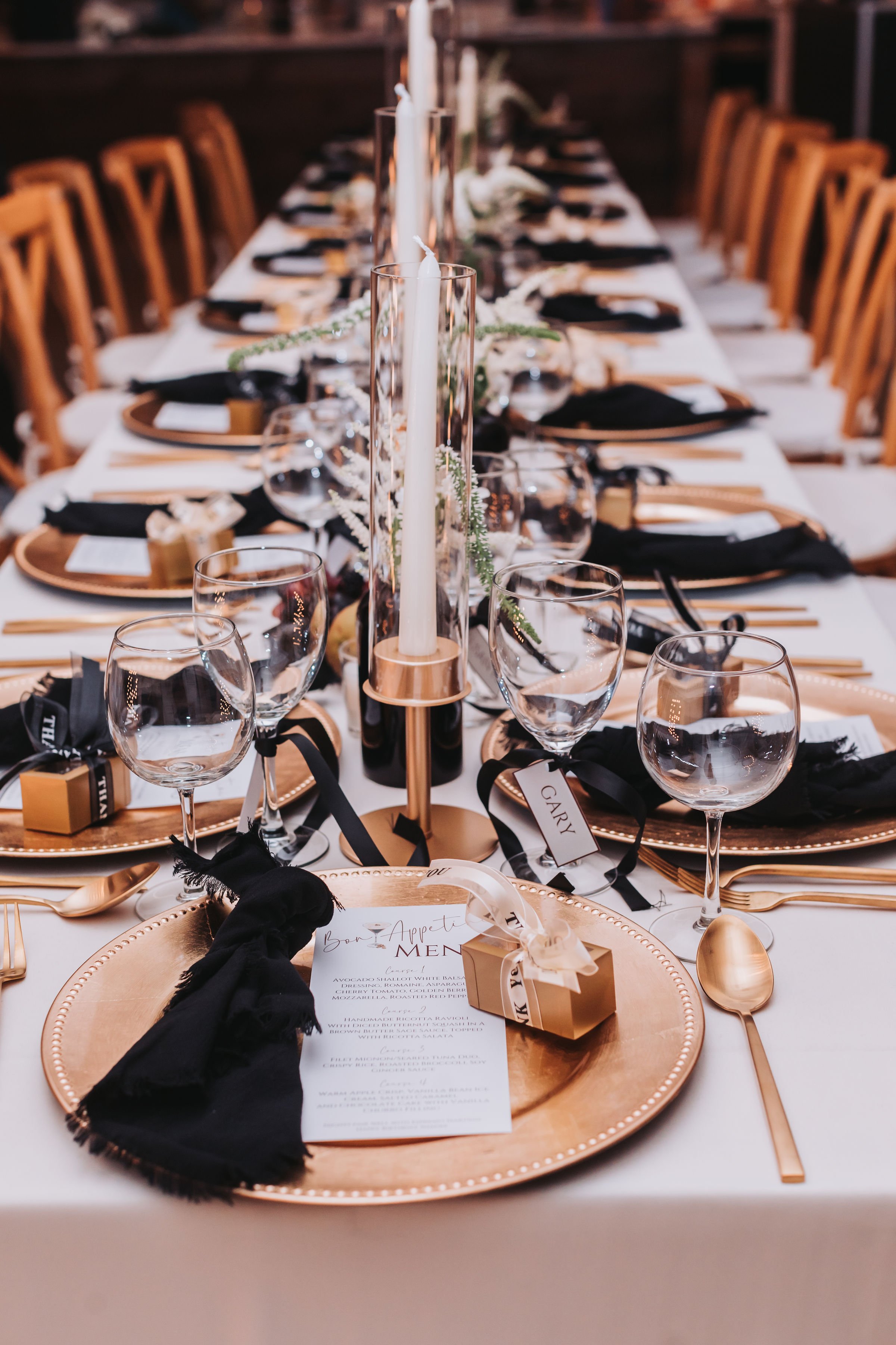 A long dinner table set for a formal event with gold, black, and white decor. The table has gold chargers, black napkins, and clear wine glasses. Tall candles and greenery serve as centerpieces. Personalized menu cards and small favor boxes are on each place setting.