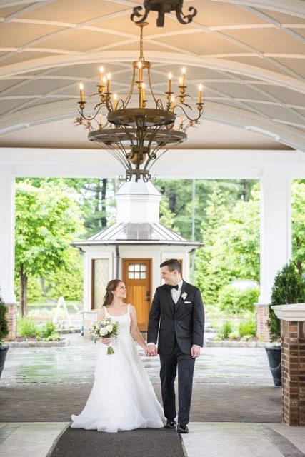 Bride and groom in wedding attire standing in front of a white vintage car with a historic building and lush greenery in the background.