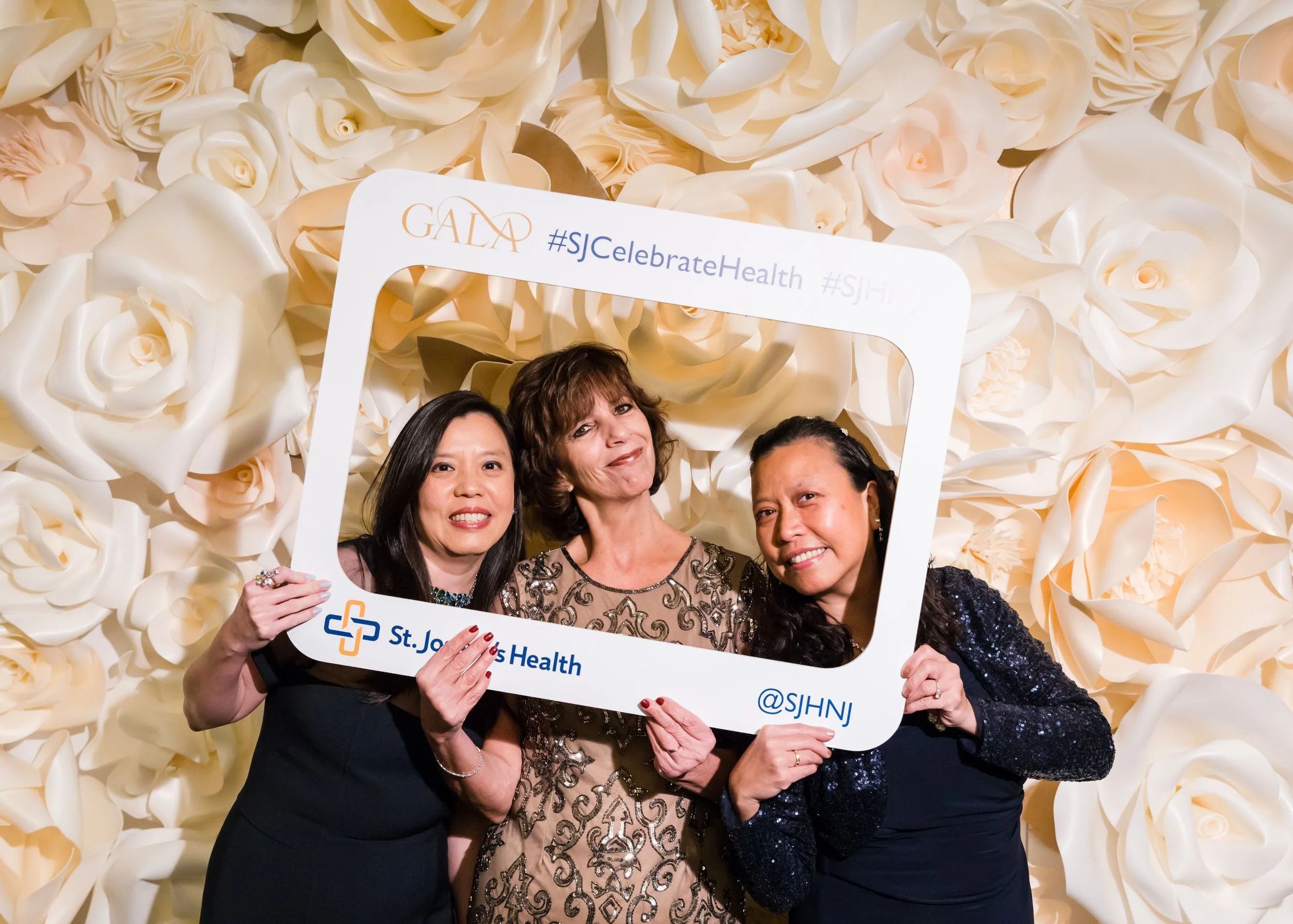 Three women holding a photo frame at a health event with a backdrop of cream-colored roses. The frame has hashtags #SJCelebrateHealth and #SJHN and the logo of St. John's Health.