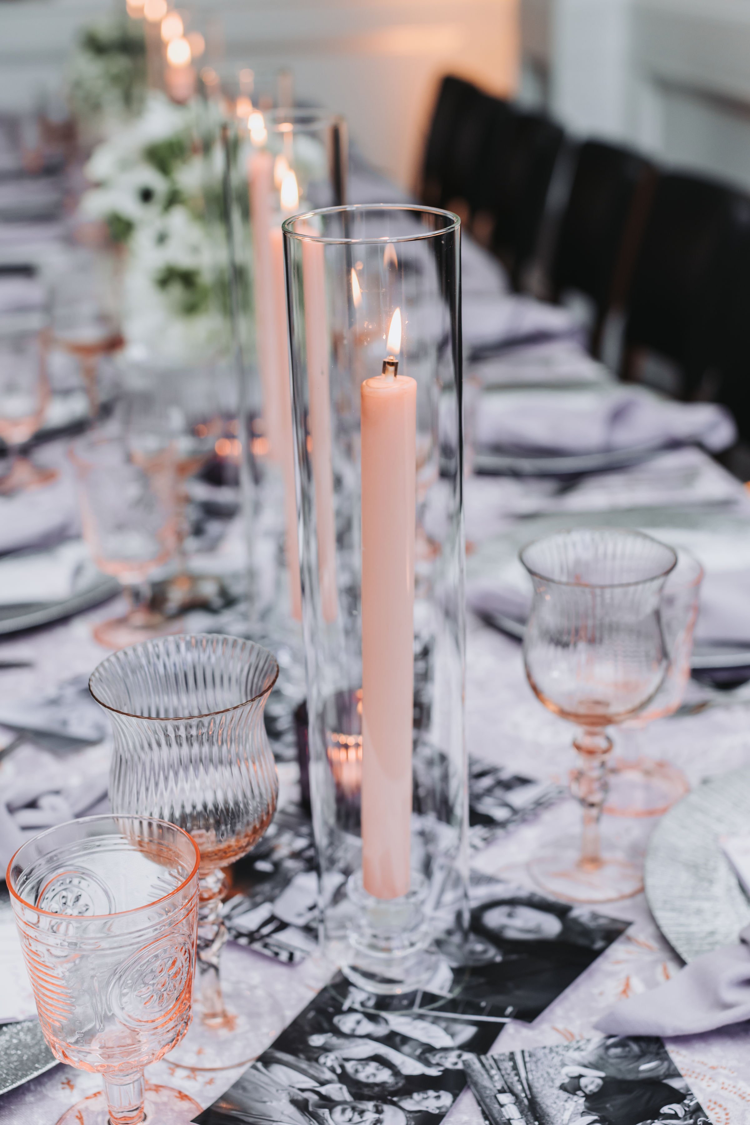 A table setting with pink-rimmed glassware, a tall candle in a glass holder, and a blurred background featuring a bouquet of white flowers and a series of lit candles.