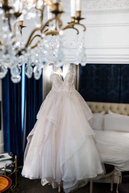 Elegant white wedding dress hanging in a luxurious room with a chandelier and dark curtains.