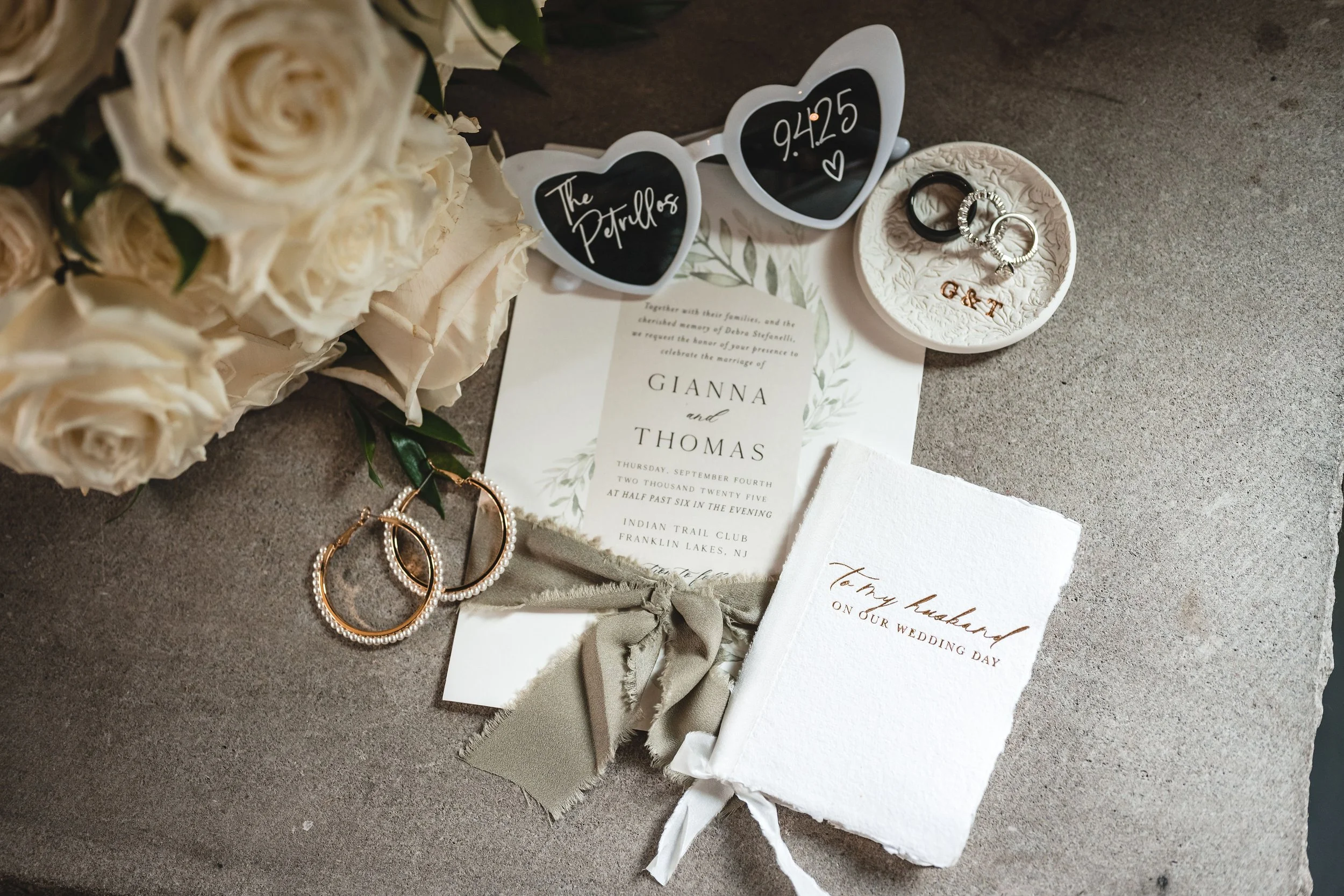 Wedding invitation suite, including a floral card with the names Gianna and Thomas, a white envelope with handwritten text, a pair of pearl and gold wedding rings, and a pair of heart-shaped sunglasses with the time 9:45:25 written on one lens.