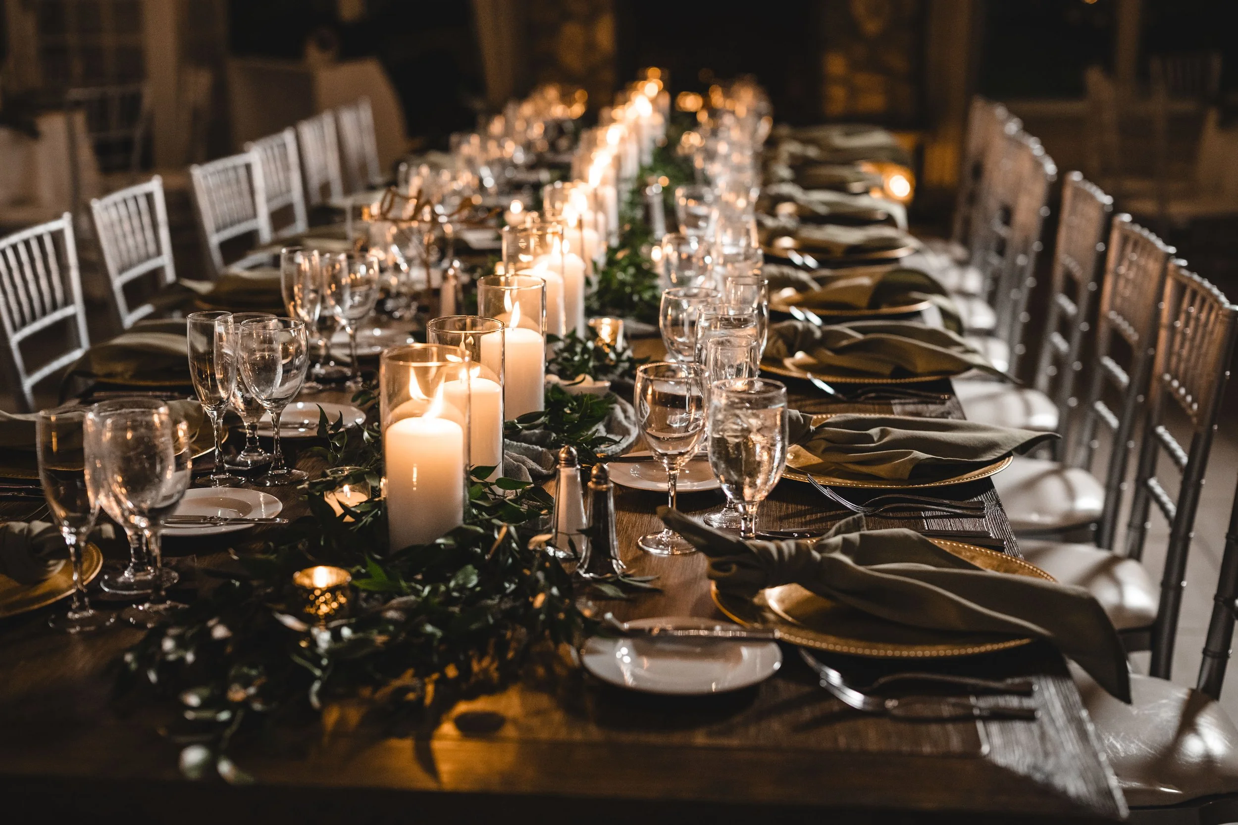 A long dining table set for an event, decorated with candles, greenery, glassware, and plates, in a warmly lit room.