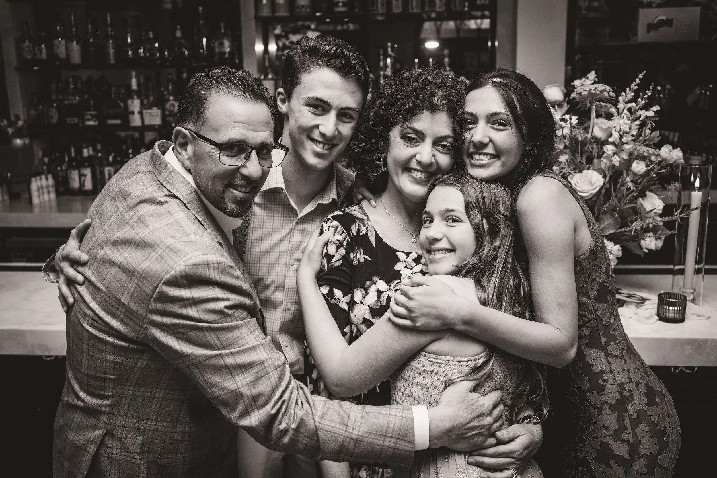 A group of six people, including three adults and three children, are hugging and smiling together, in an indoor setting with bottles and flowers in the background.