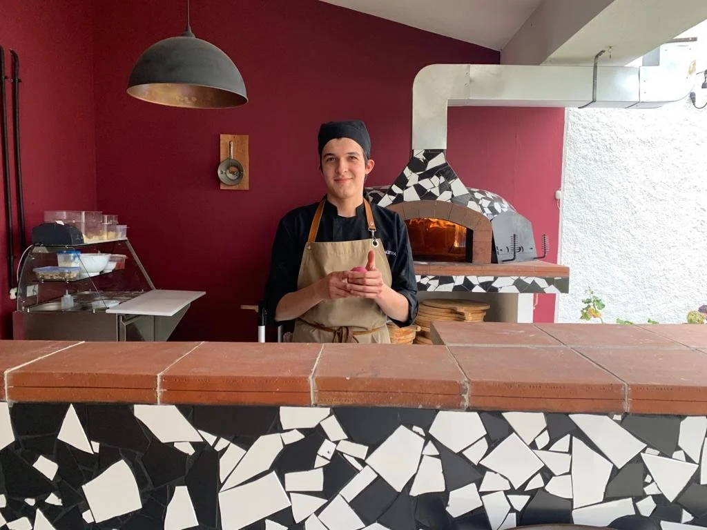 Young male chef (Guillo) standing behind a counter in front of a pizza oven, wearing a black chef coat and beige apron, with a black chef hat, in a kitchen with a maroon wall and black and white tile decor.