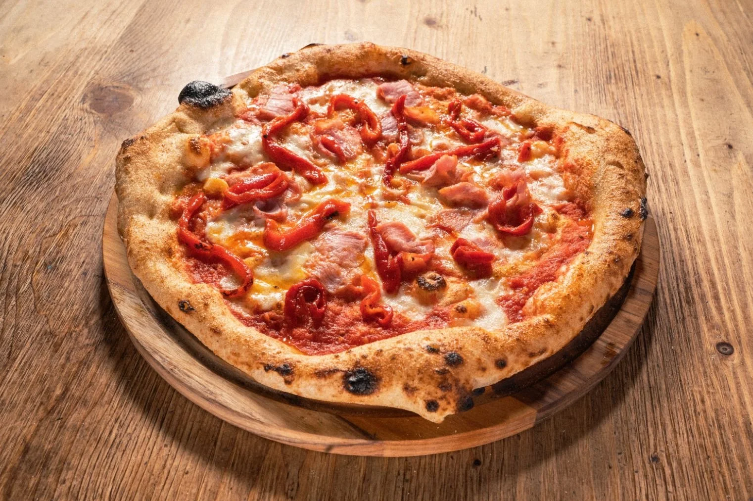 Our signature pizza (La Española) with mozzarella, red bell peppers, tomato sauce, and bacon on a wooden board, on a wooden table.