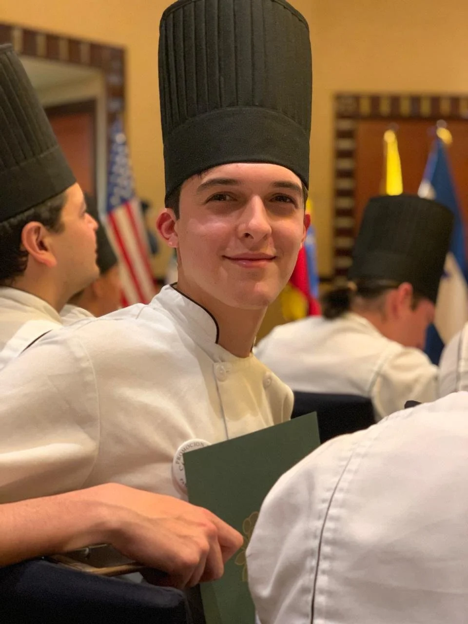 Young male chef (Guillo) wearing a tall black chef hat and white chef uniform, sitting at a graduation with other chefs in a formal setting with various national flags in the background.