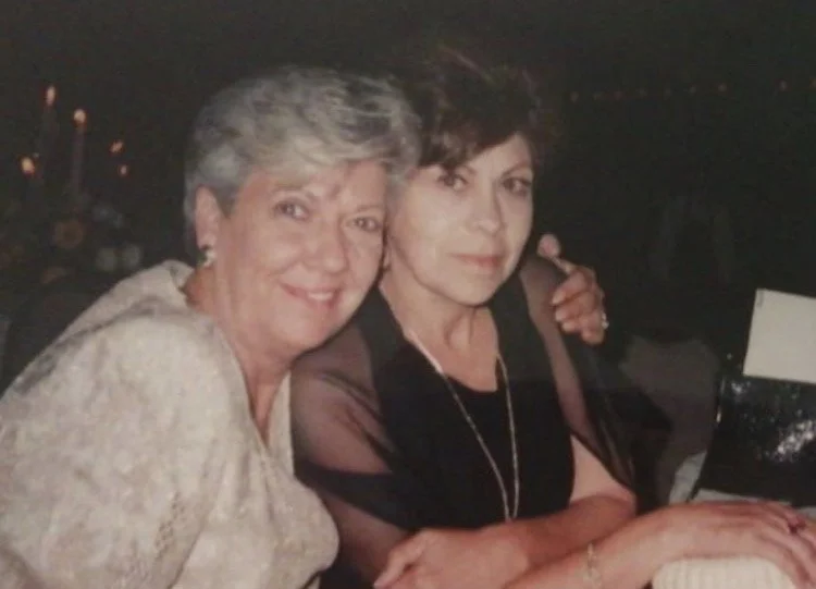 Two women (Carmen and Lily) sitting closely together at a table, smiling at the camera. One has short gray hair, and the other has short dark hair. They appear to be at a social gathering or event, with a dark background and candles visible. Vintage.