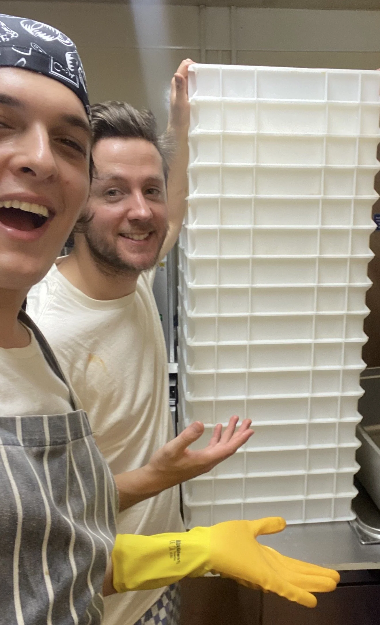 Two men (Guillo and Oscar) smiling in a kitchen, one wearing a striped apron and yellow gloves, the other holding a pile of large plastic trays.