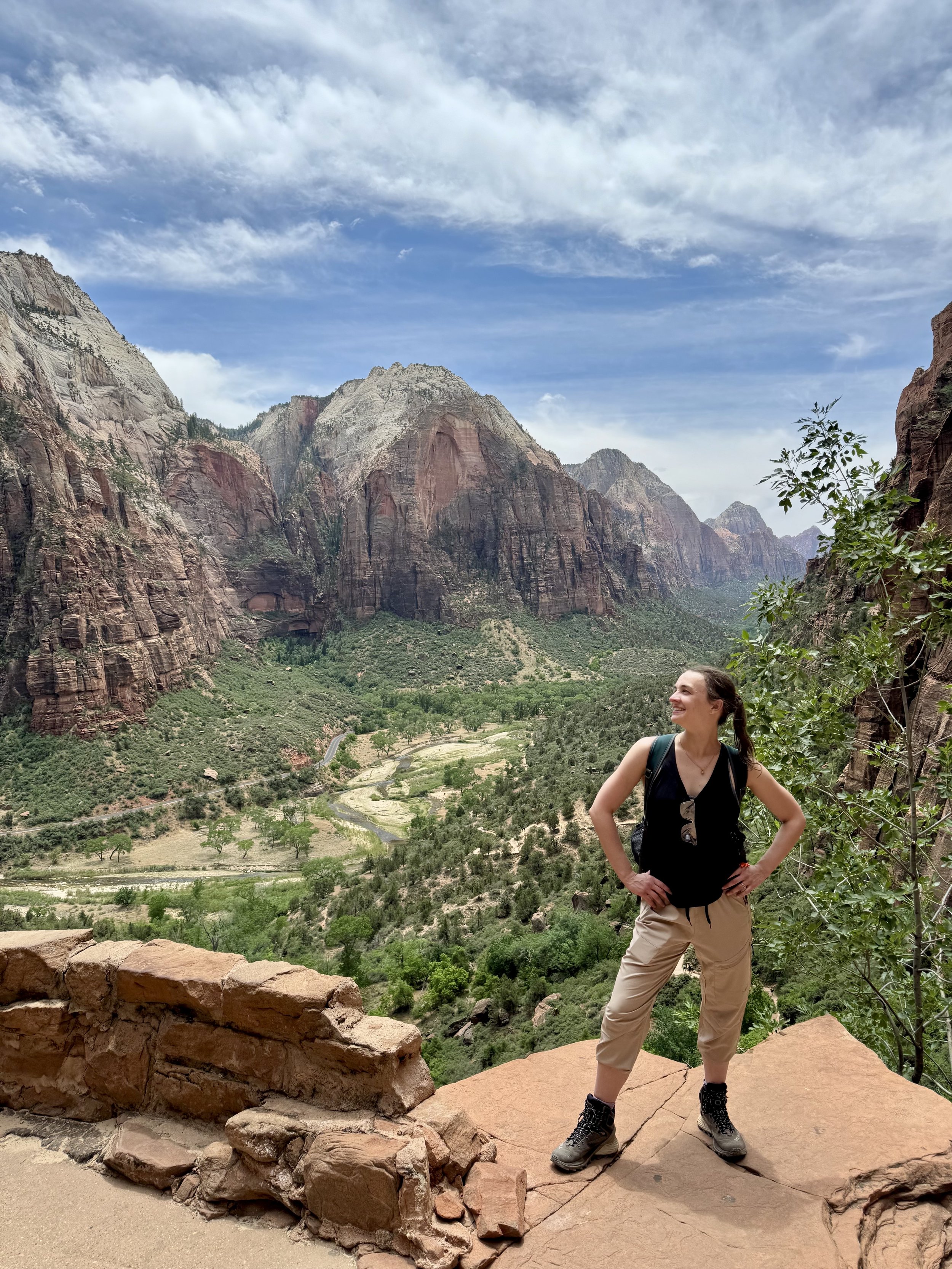A woman standing on a rock ledge in Zion National Park with a view of canyon walls and a valley, smiling and looking to her right, wearing beige pants, a black top, hiking boots, and carrying a backpack.