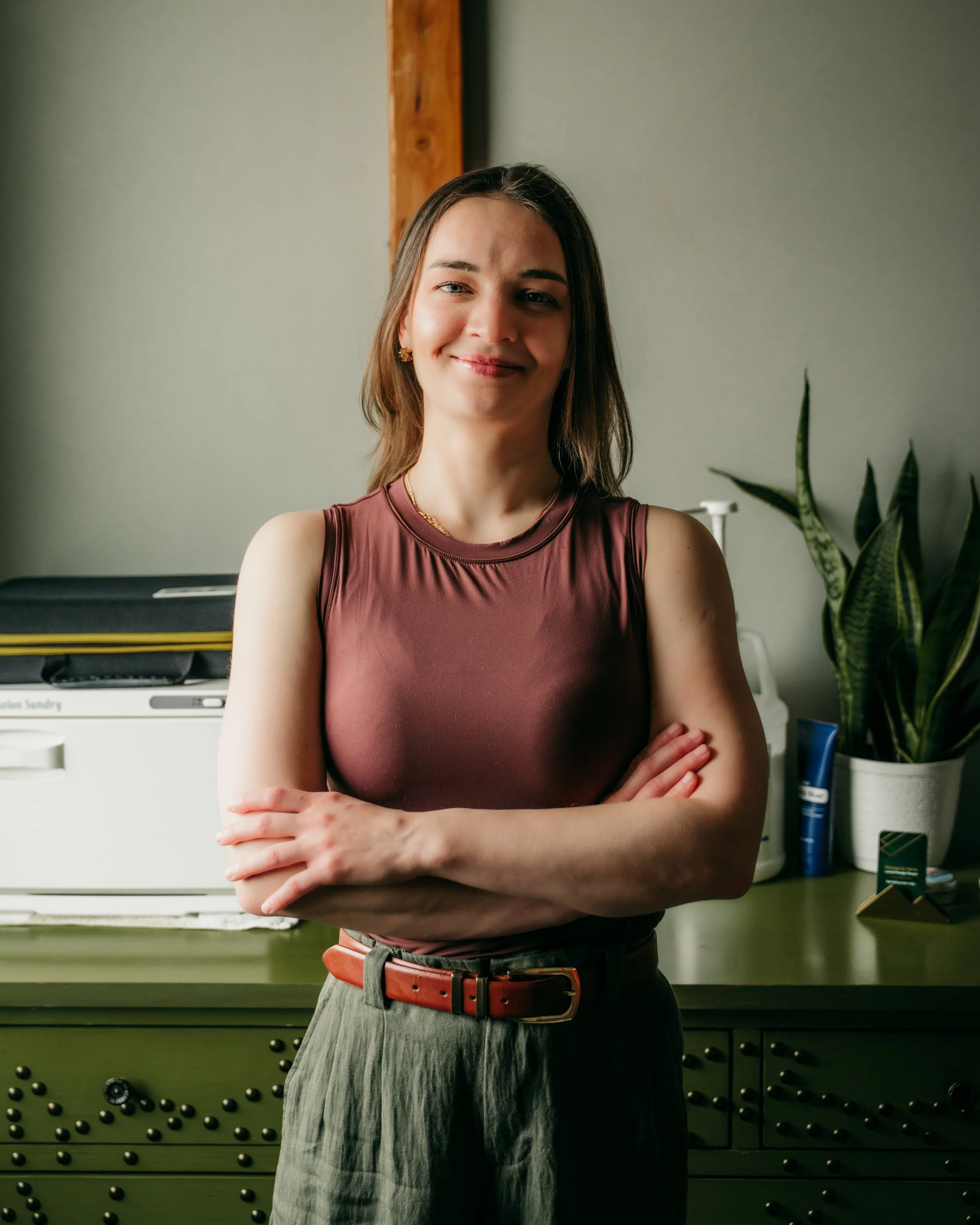 A woman with brown hair, wearing a sleeveless burgundy top and gray pleated pants, standing with arms crossed in front of a dark green cabinet and a potted snake plant. She has earrings, a necklace, and a red belt.