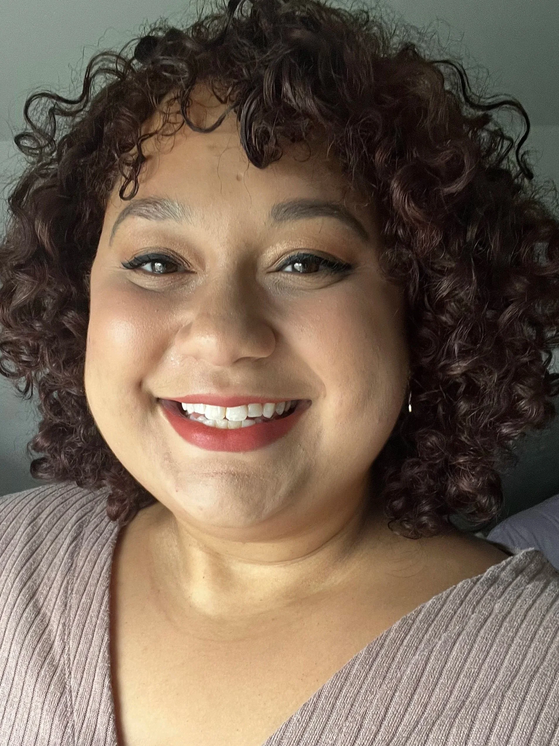 Close-up of a woman smiling, wearing makeup, and a ribbed gray top.