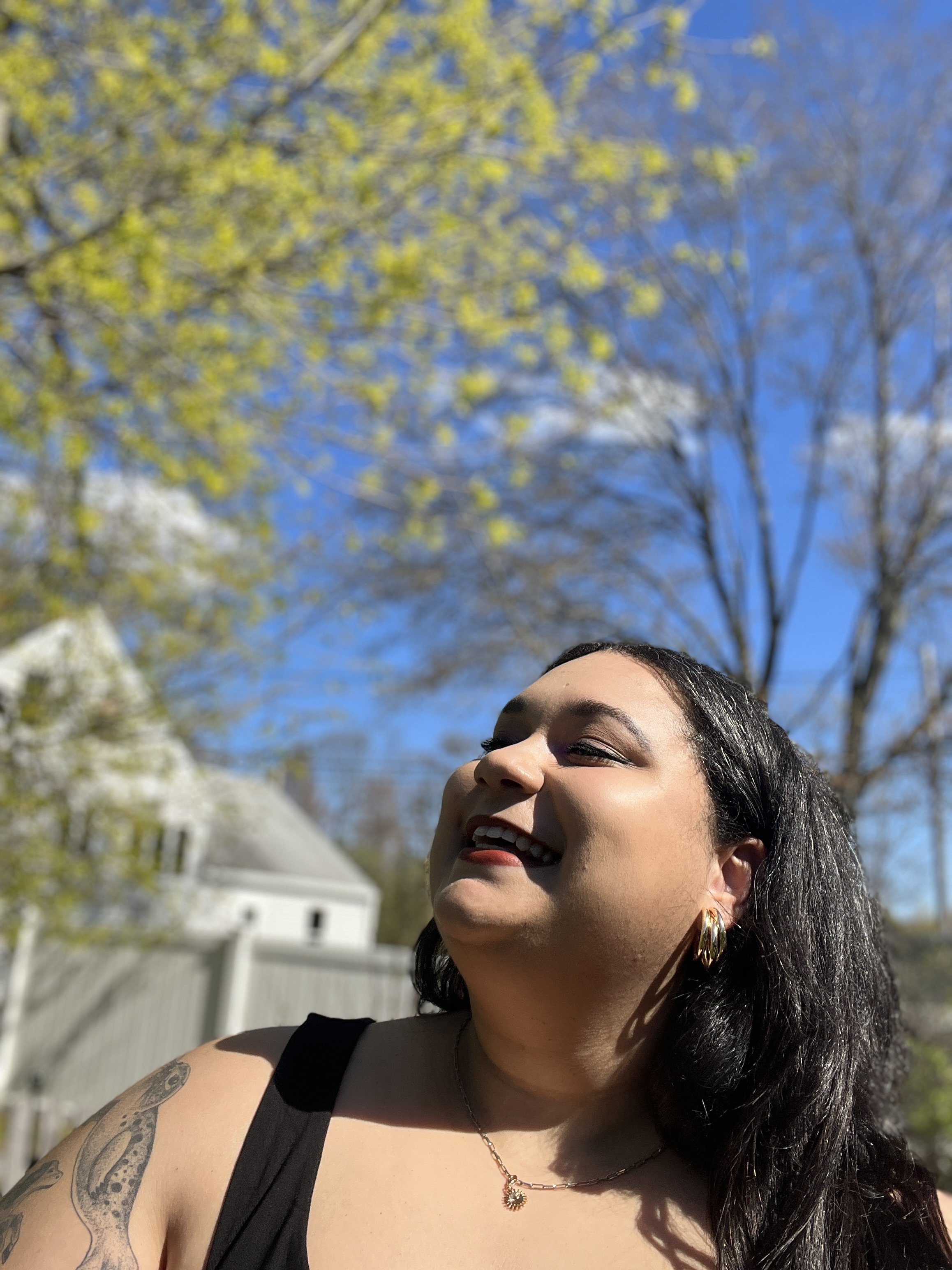 A woman with long dark hair, wearing a black top, gold hoop earrings, a necklace with a sunflower pendant, and tattoos on her arm, smiling outdoors with a clear blue sky, yellow and brown trees, and a white house in the background.