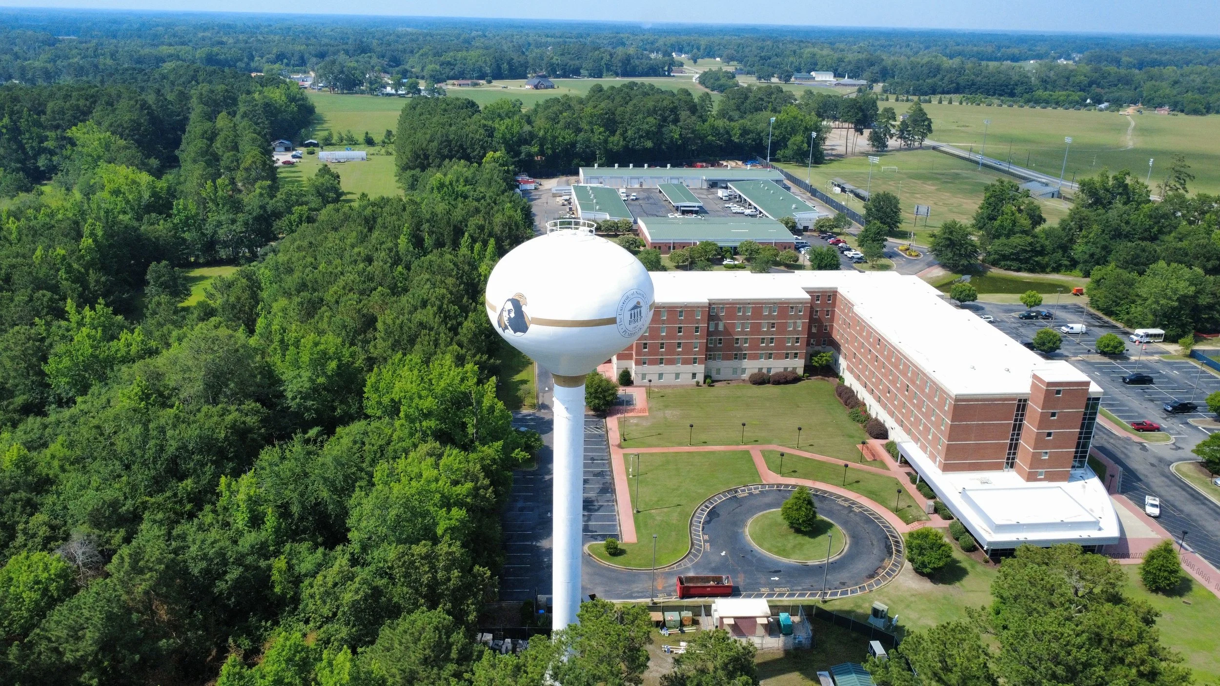 Aerial view of a university campus with a water tower featuring a logo and a person's profile, surrounded by trees and sports fields.