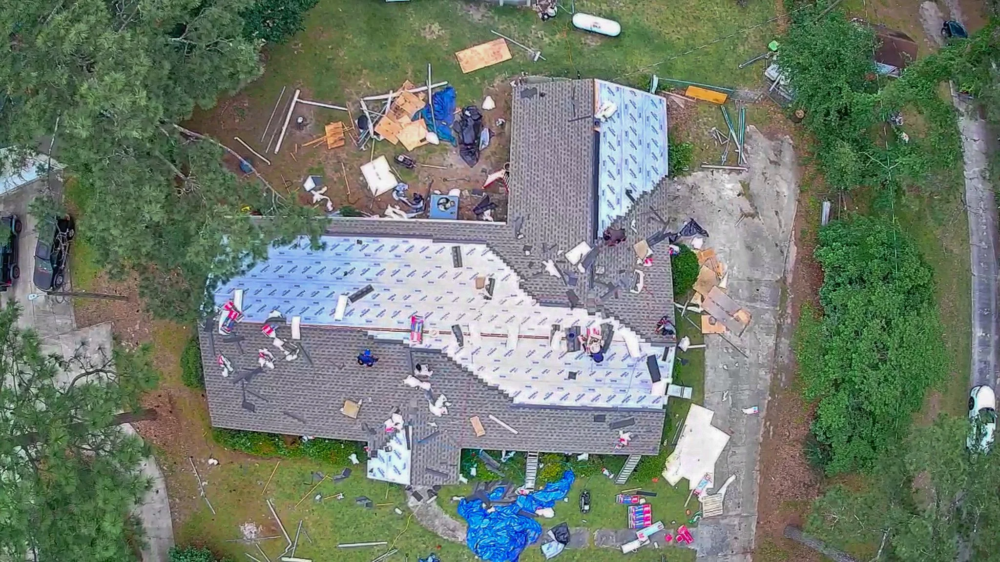 Aerial view of a house under construction with tools, building materials, and equipment scattered around the yard, surrounded by trees and a driveway with parked cars.