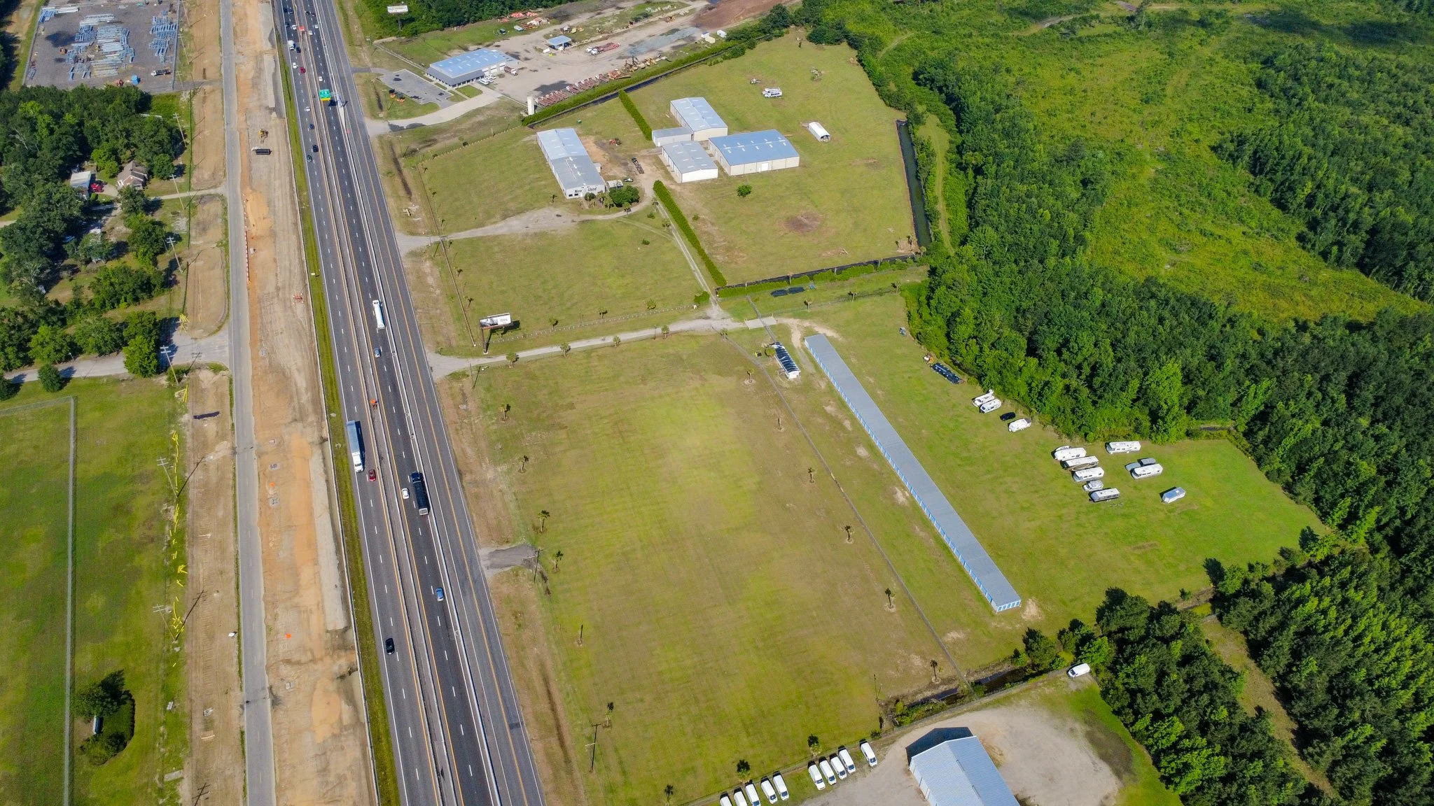 An aerial view of a highway running parallel to a fenced grassy area with scattered small buildings and parking lots, surrounded by dense green trees.