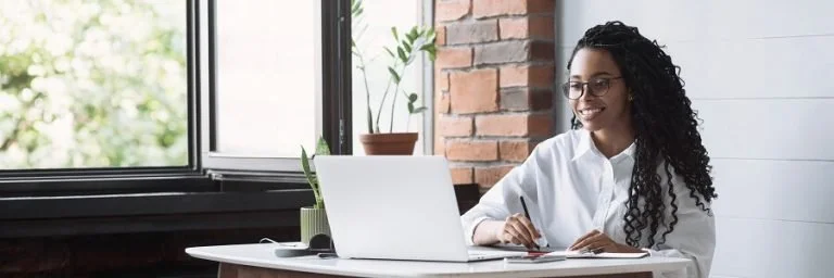 A woman with curly hair and glasses working on a laptop at a desk in a bright room with a large window.
