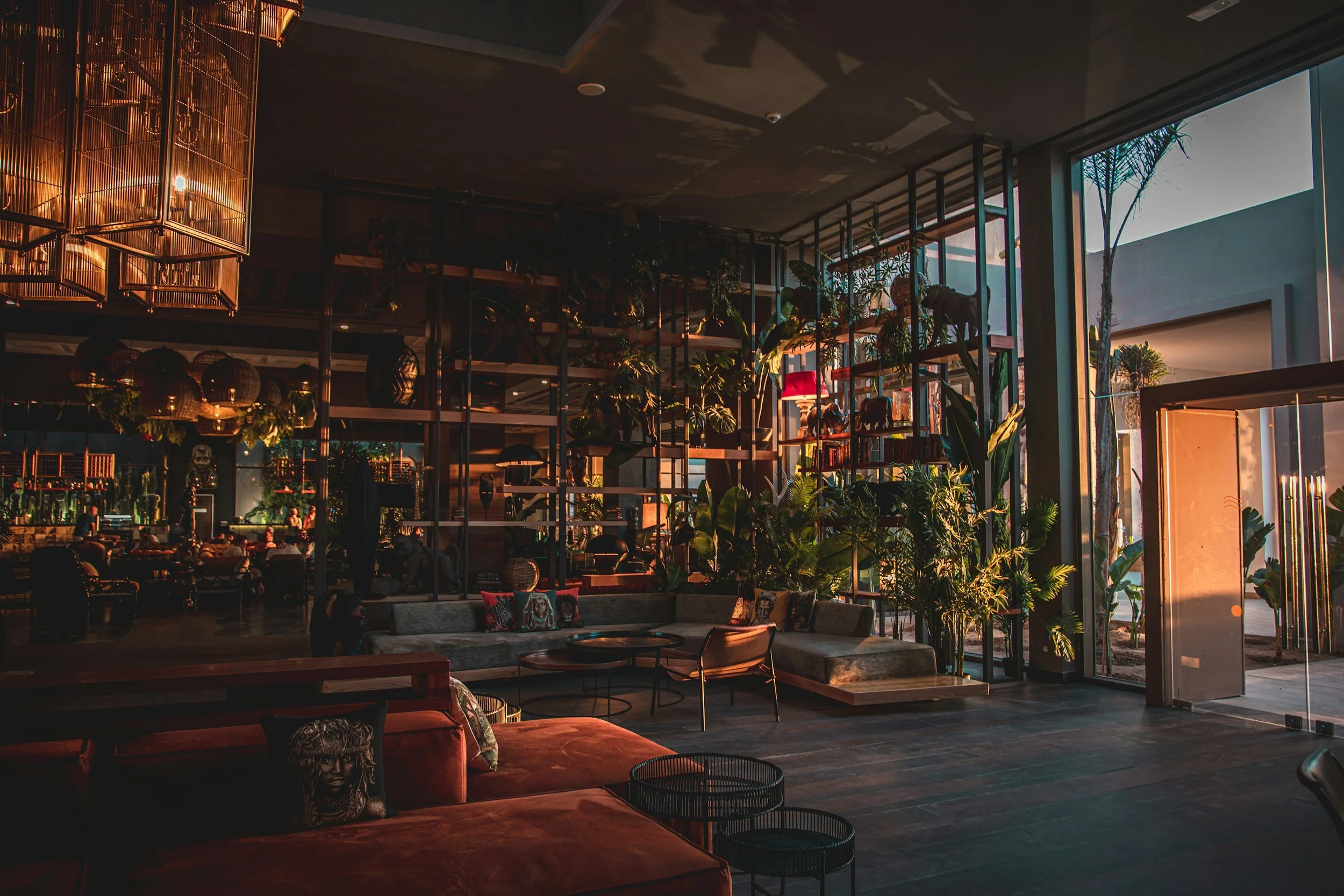 Interior of a modern lounge or hotel lobby with large windows, abundant indoor plants, a gray sofa with decorative pillows, warm ambient lighting, and mezzanine levels with additional greenery, creating a cozy atmosphere.