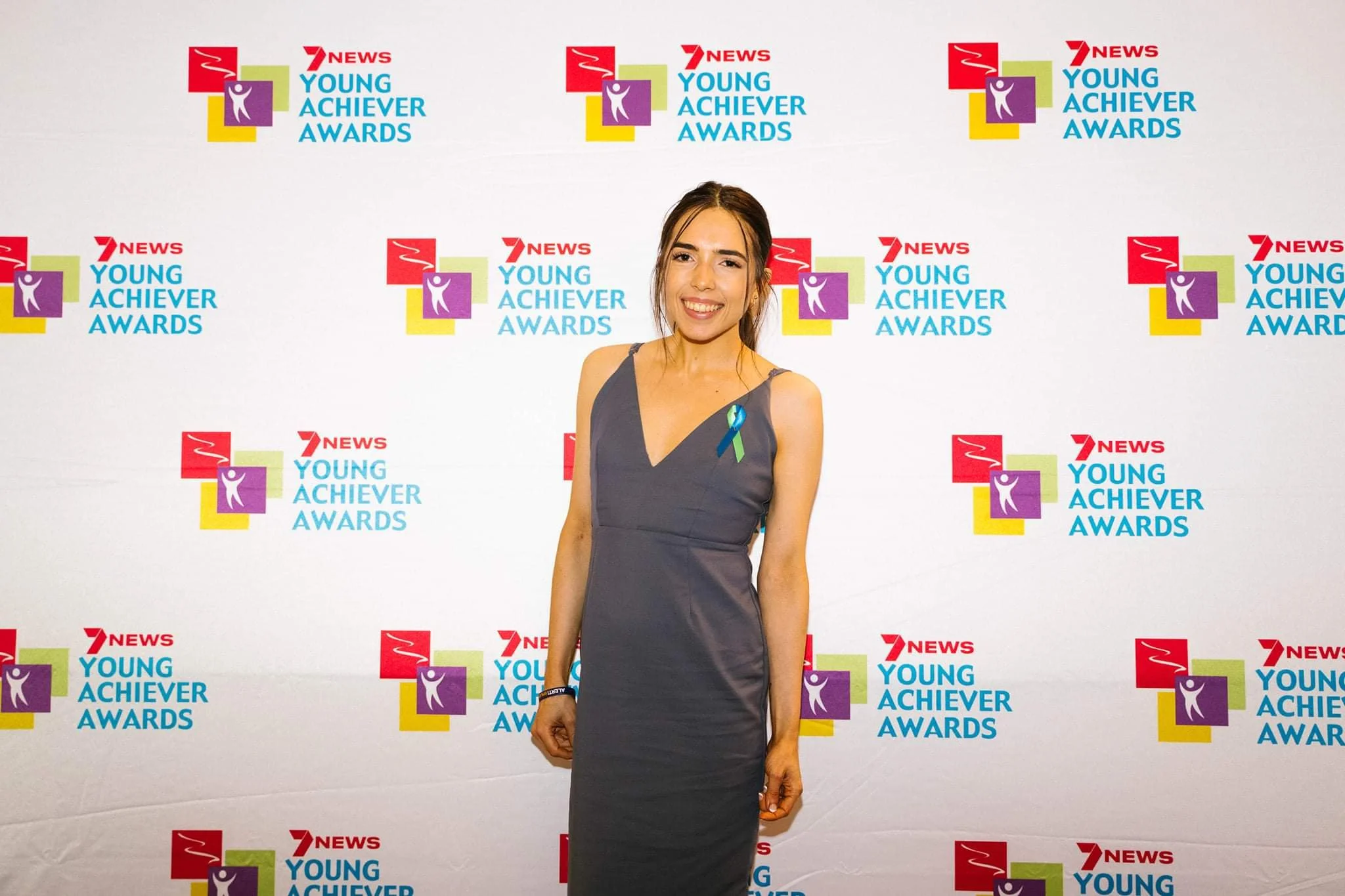A woman smiling at an awards event with a backdrop displaying the Young Achiever Awards logo and 7 News logo.
