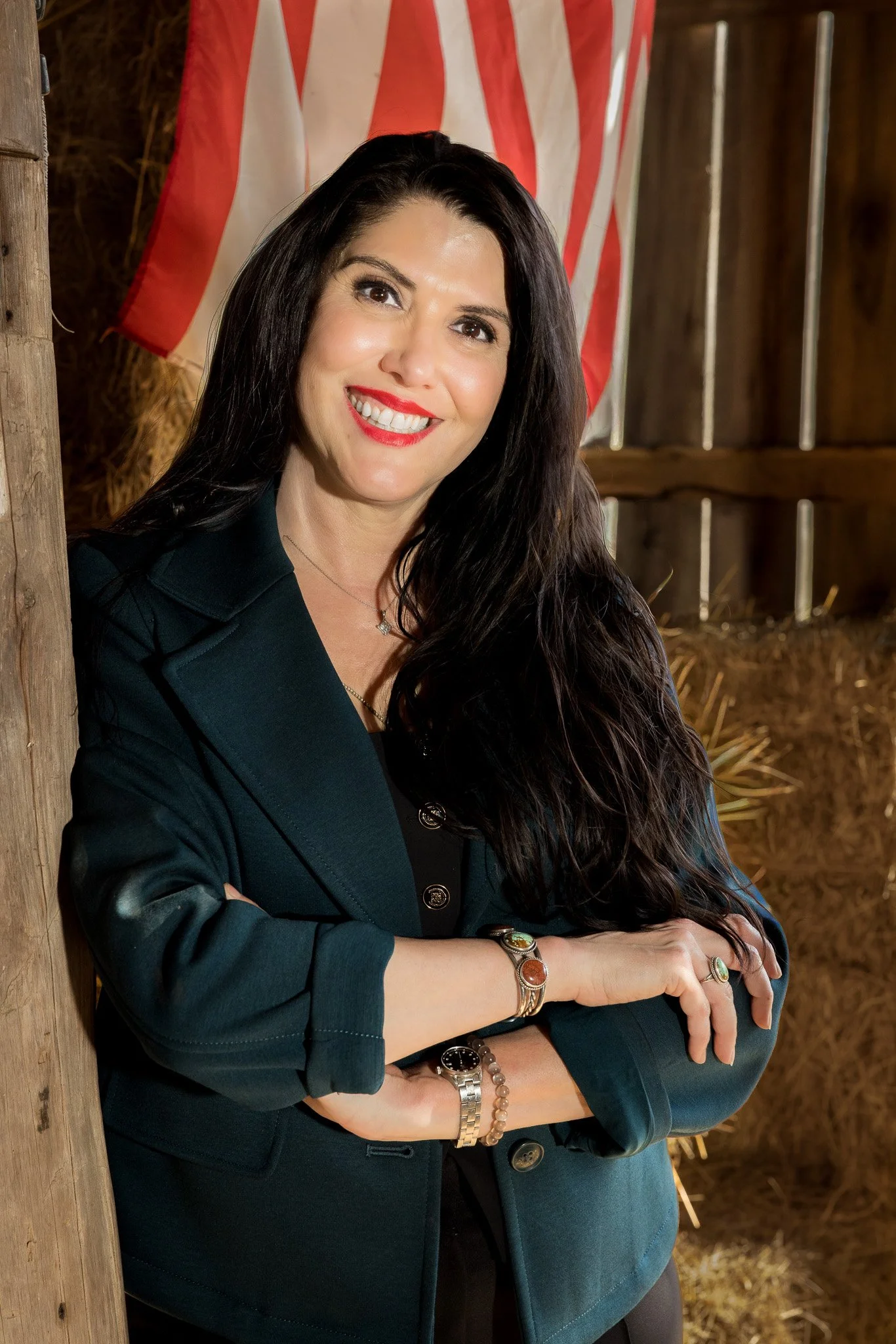A woman with long dark hair and red lipstick standing in a barn with hay bales, crossing her arms and smiling. Wilson County Mayor Candidate, Democrat, Democratic, Running, military, veteran, Lebanon, Mt. Juliet, Mount Juliet, mt juliet, watertown