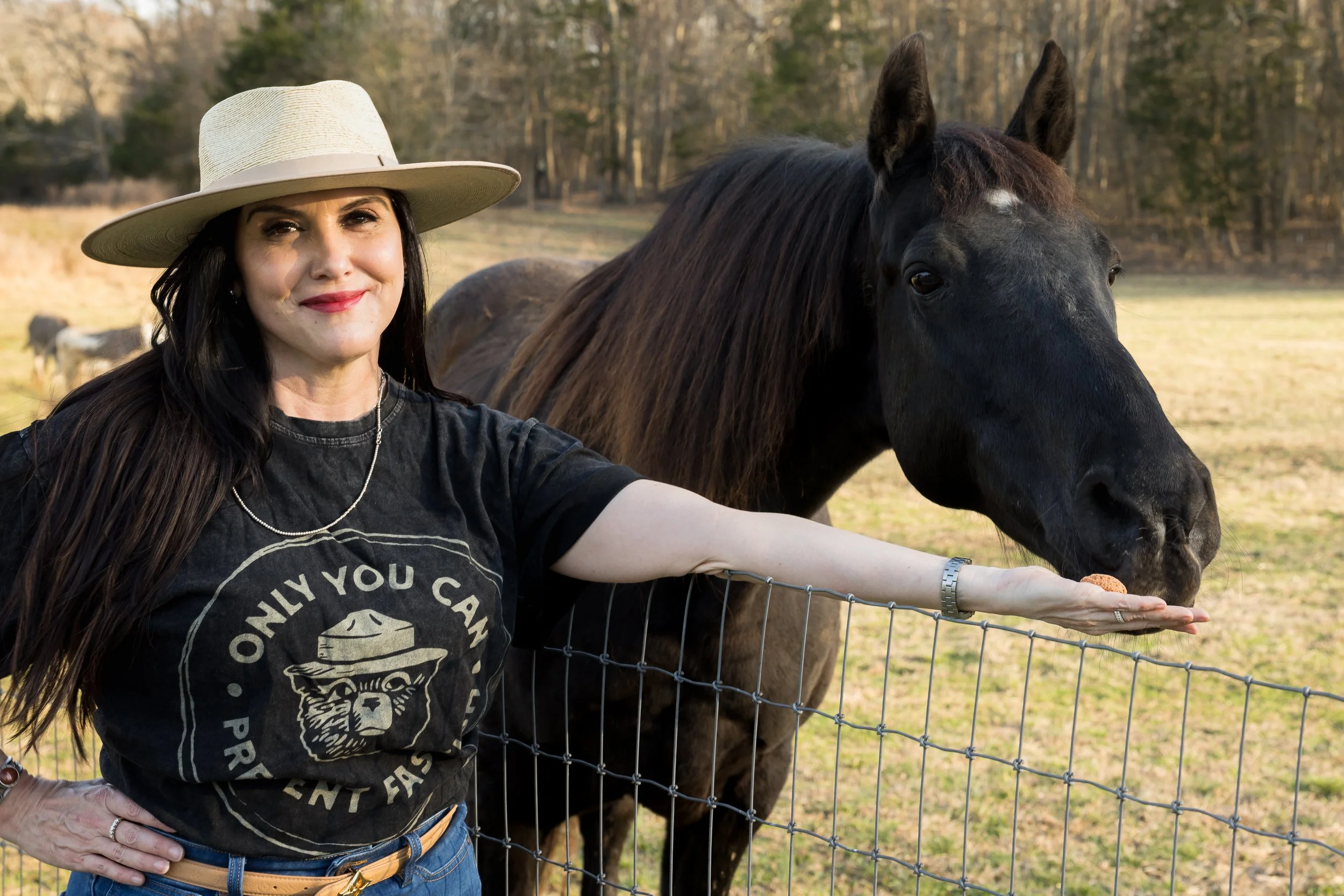 A woman with long dark hair wearing a wide-brimmed hat, a black t-shirt with a graphic and text, standing outdoors next to a black horse behind a wire fence, with a background of trees and open field.