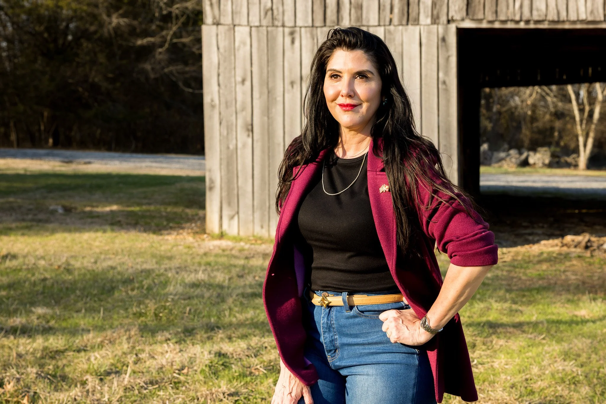 A woman standing outdoors near a wooden structure, wearing a maroon blazer, black shirt, jeans, and a star-shaped belt buckle, with trees in the background. Christina Puza, Redbird Farms, 25-acre lebanon