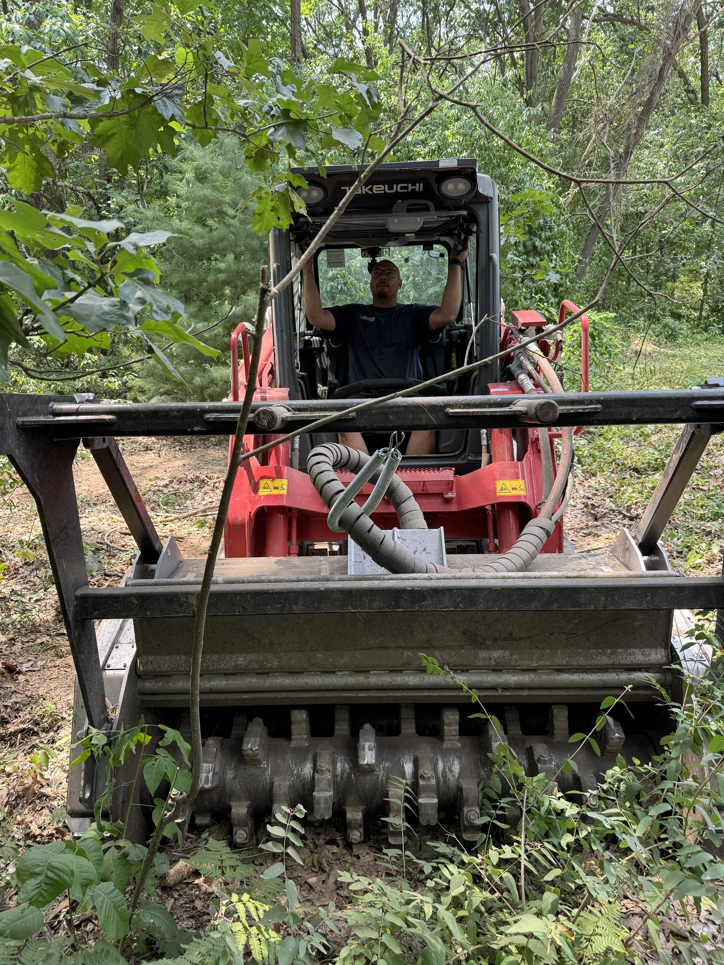 A man operating a red compact excavator in a wooded area, surrounded by green foliage and trees.