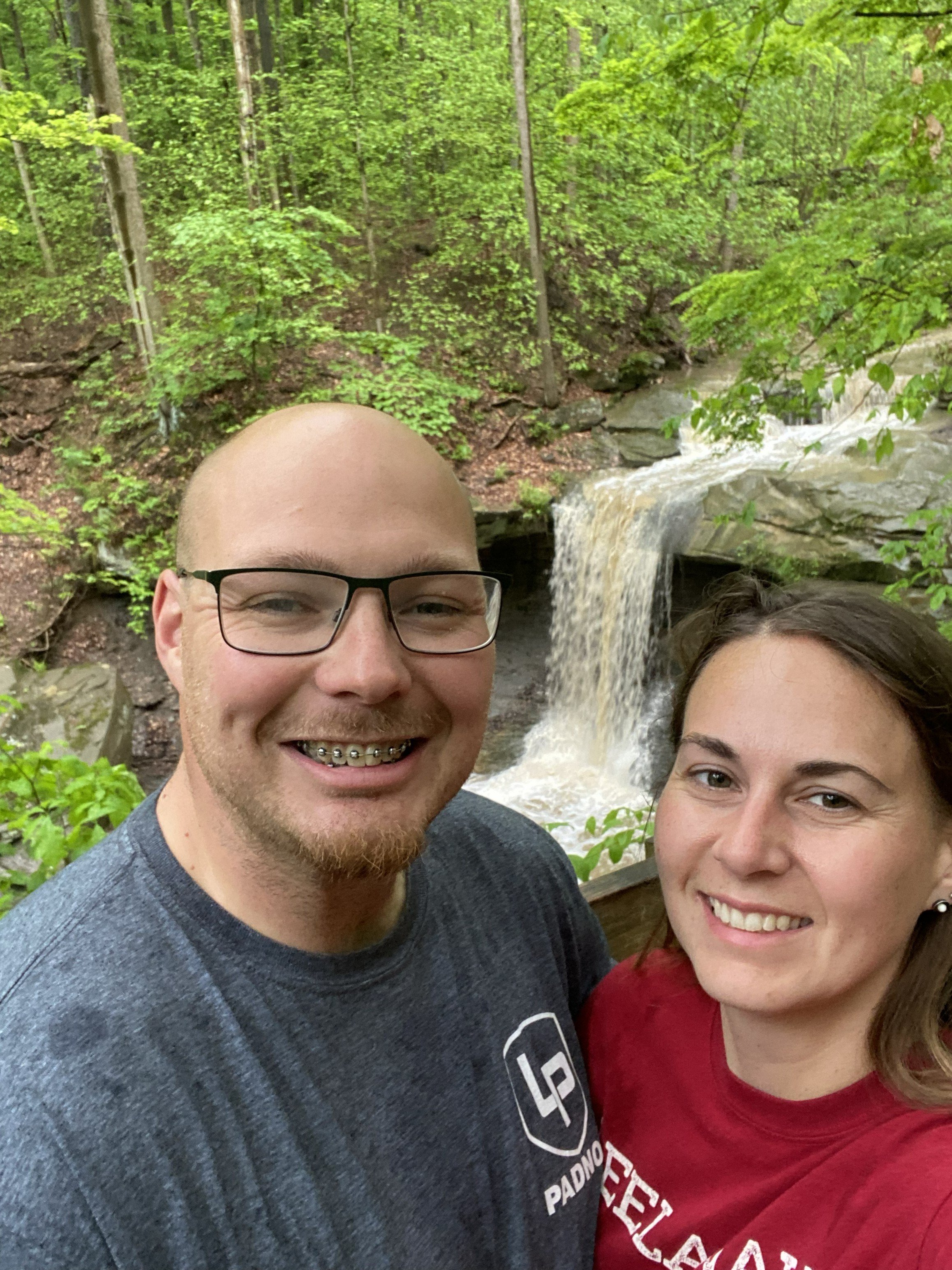 A selfie of a smiling man with glasses and braces, and a woman smiling, in front of a waterfall in a lush green forest.