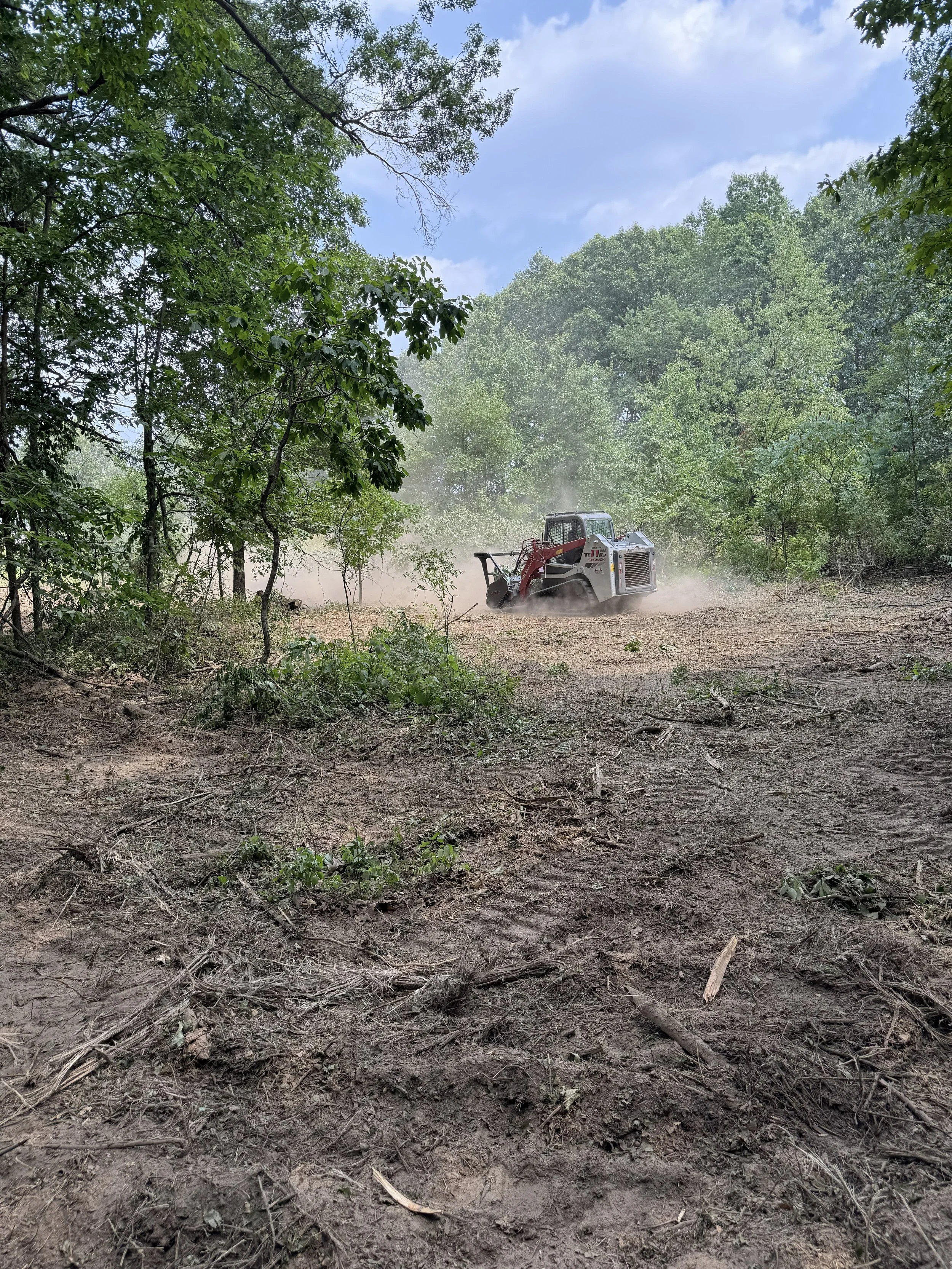 A small bulldozer working on a dirt clearing surrounded by trees, with dust rising in the air and a partly cloudy sky above.