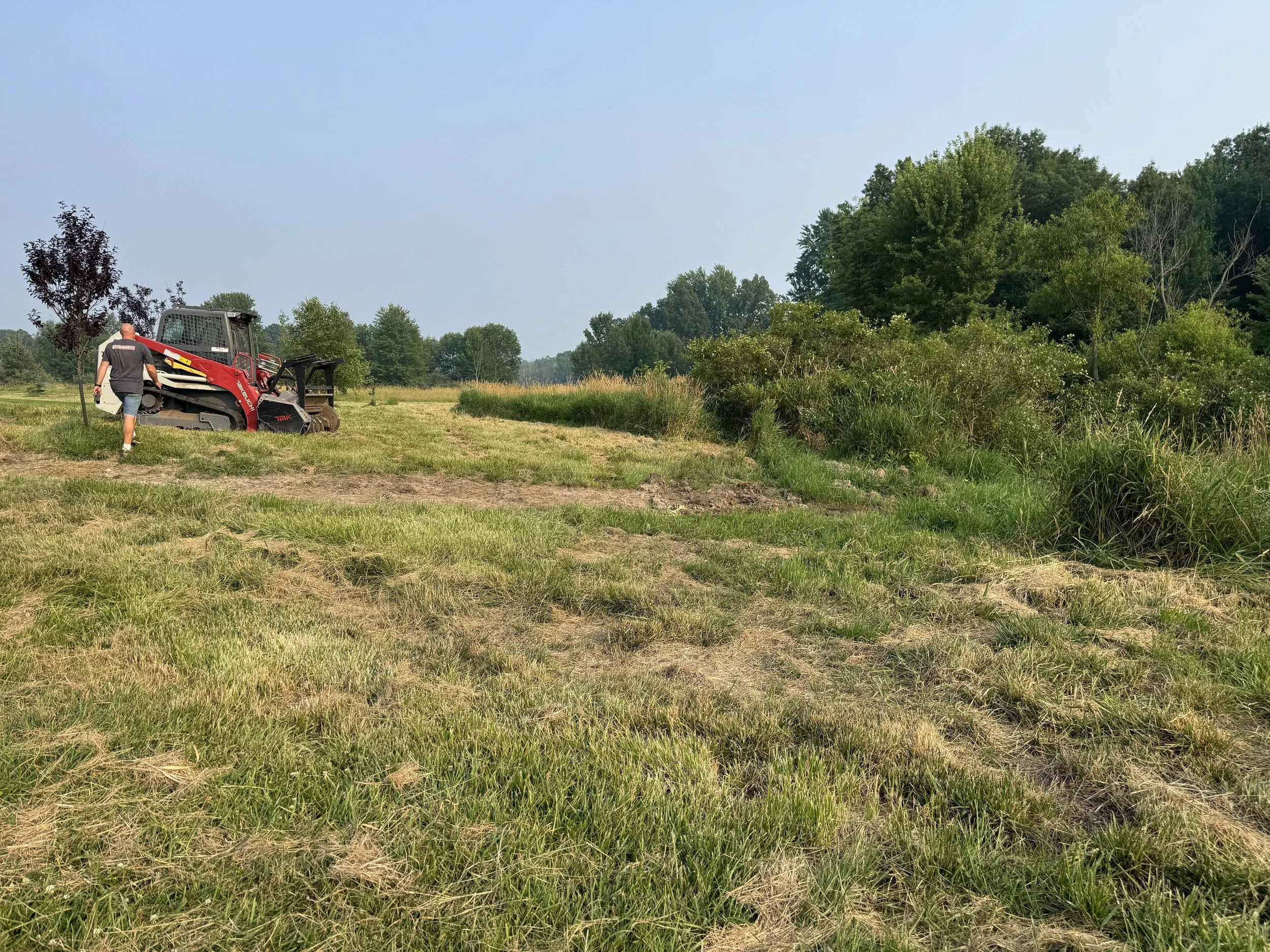 A person walking away from a red and black compact excavator in a grassy field with trees and a clear blue sky in the background.