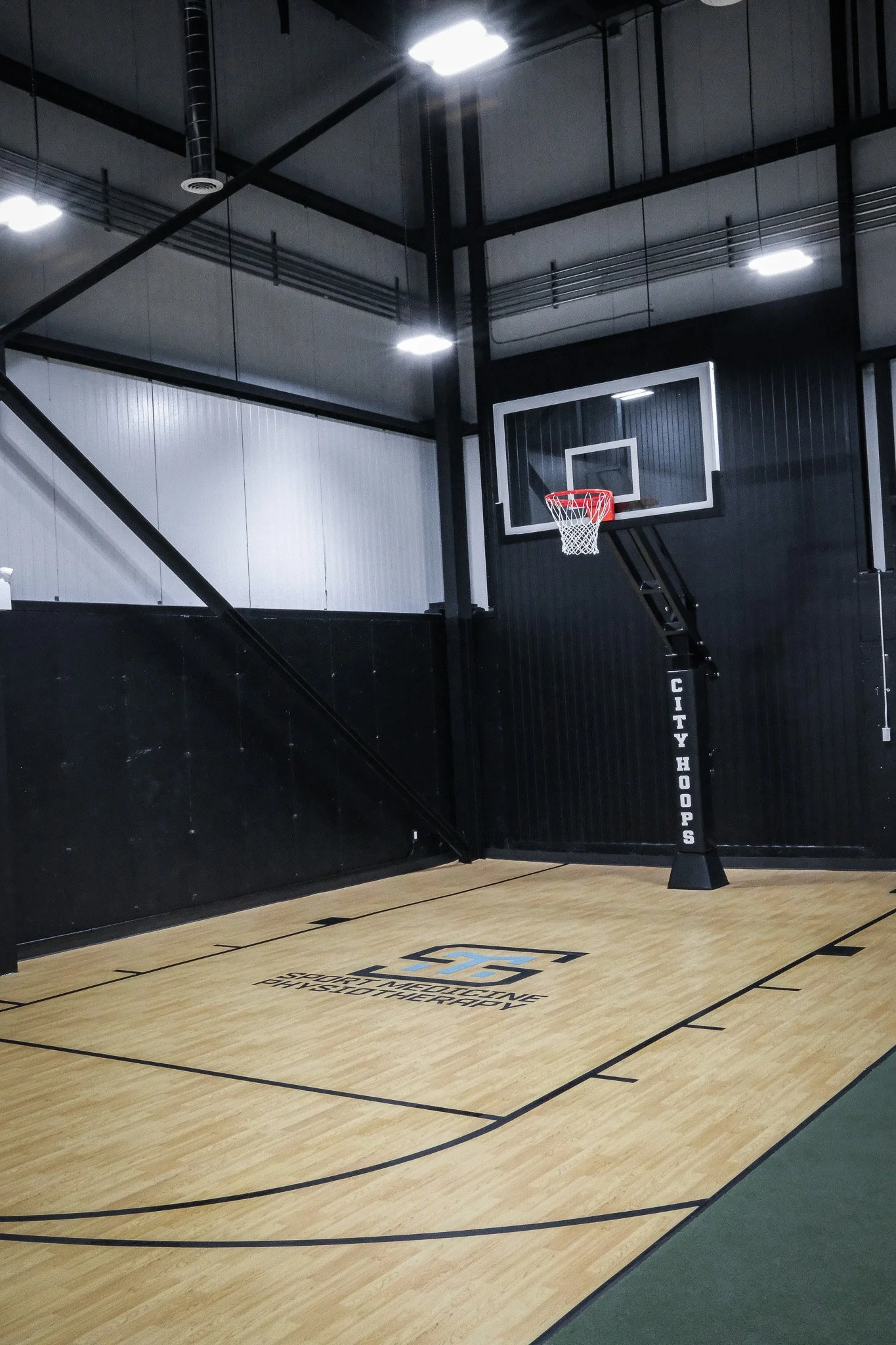 Indoor basketball court with a hoop and backboard, black walls, and a polished wooden floor with sport medicine physiotherapy branding.