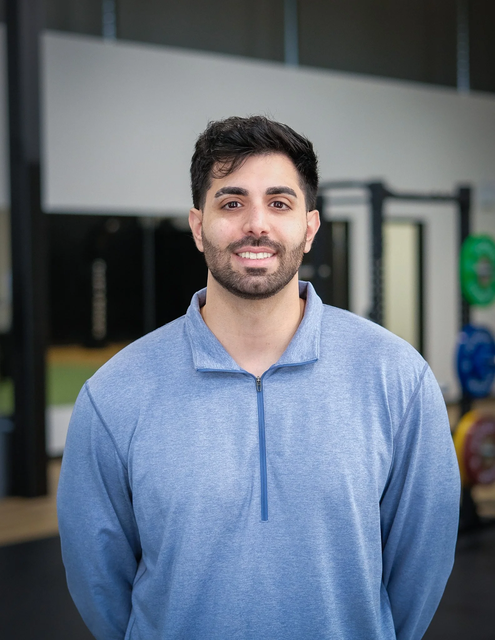 A male physiotherapist with dark hair, a beard, and a mustache, wearing a blue zip-up jacket, standing indoors with a blurred background of gym equipment.