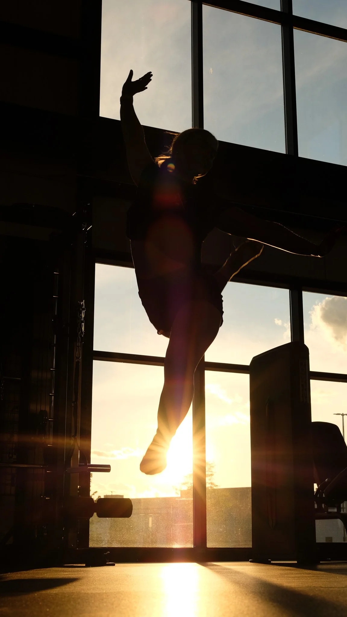 Person silhouette practicing a dance jump in a physiotherapy gym with large windows at sunset.