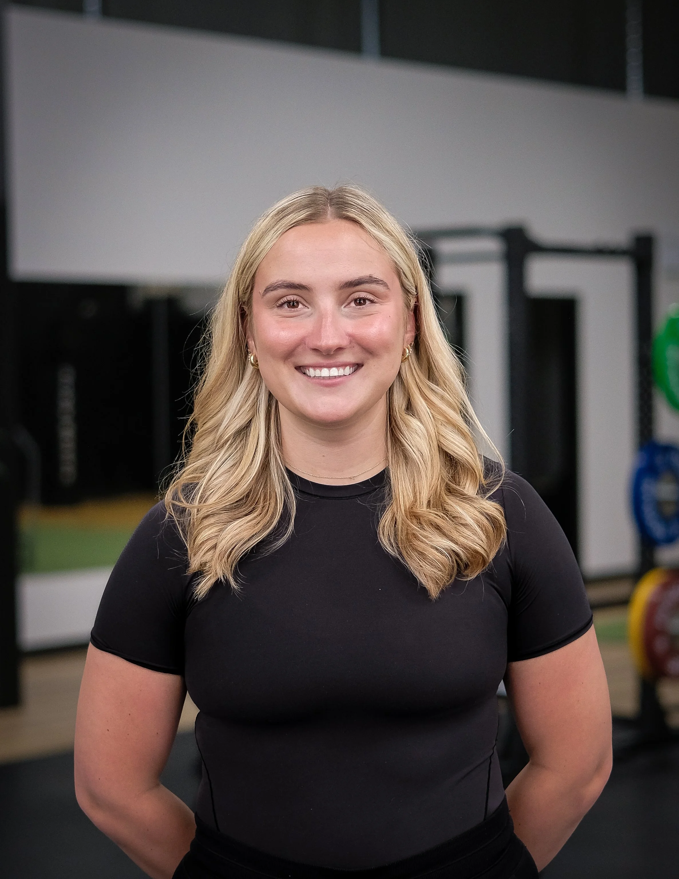 A female physiotherapist with long blonde hair, smiling, wearing a black athletic shirt, standing in a gym or fitness center.