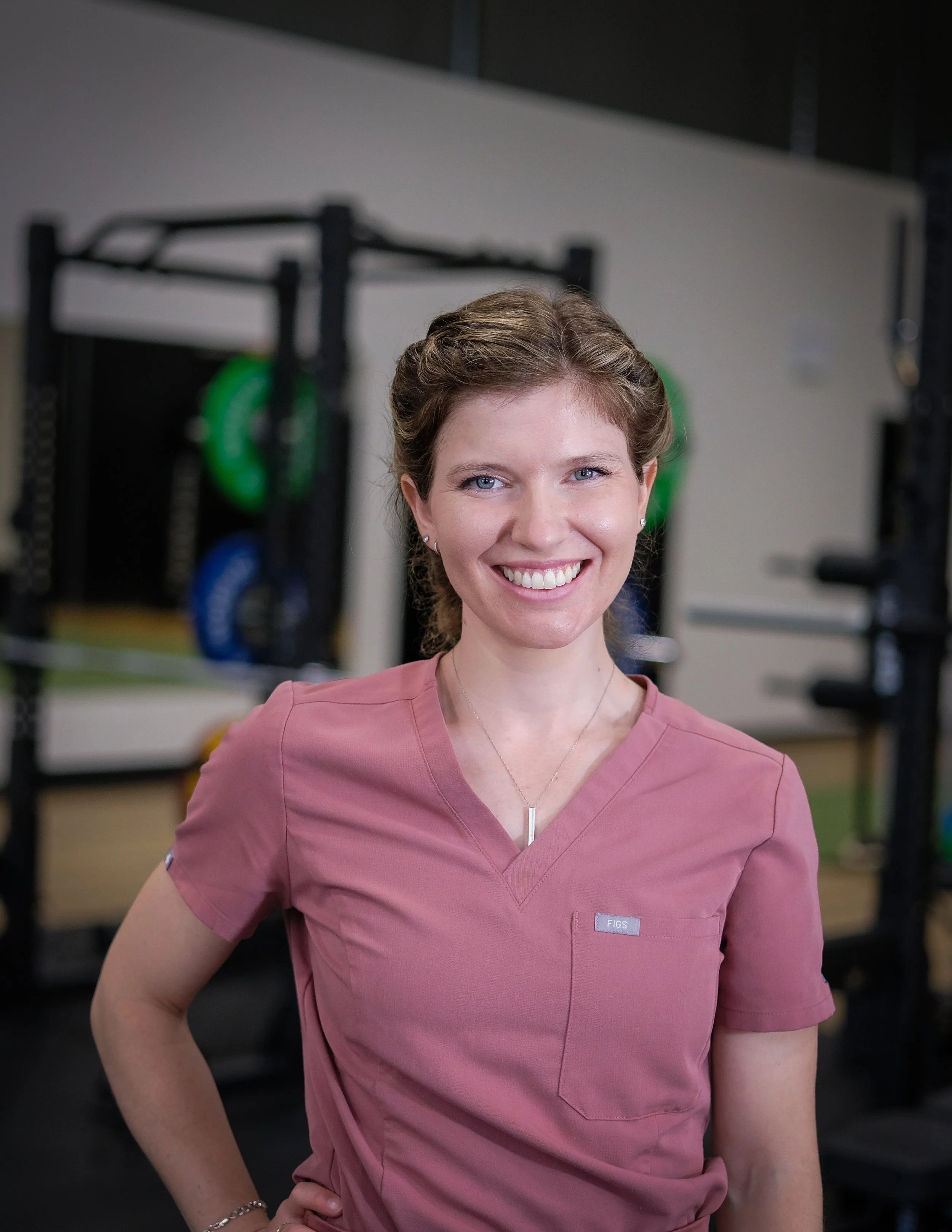 A smiling female physiotherapist in pink medical scrubs standing in a gym with black storage racks and colourful exercise weights in the background.